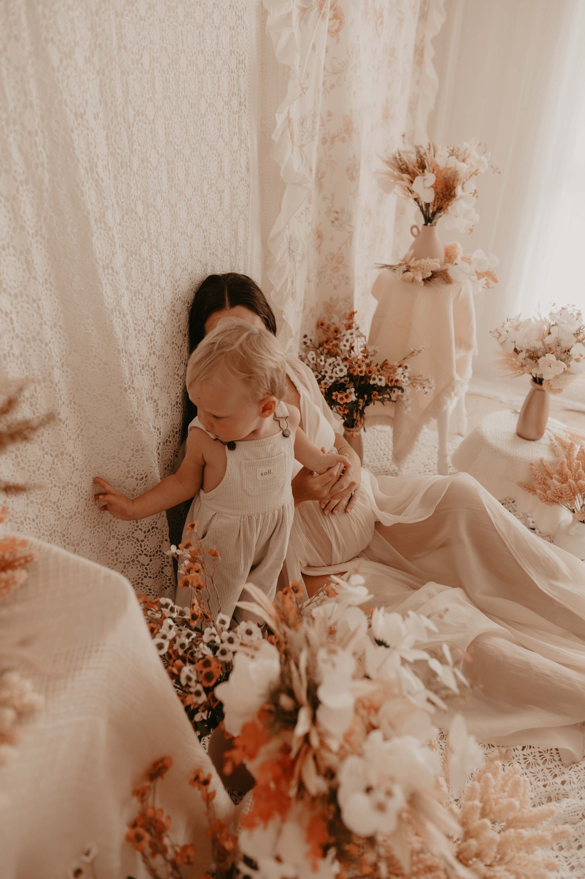 A woman and a young child sitting on a cream-colored lace-covered floor surrounded by peach, white, and pink flowers in vases, with a textured cream-colored wall in the background.