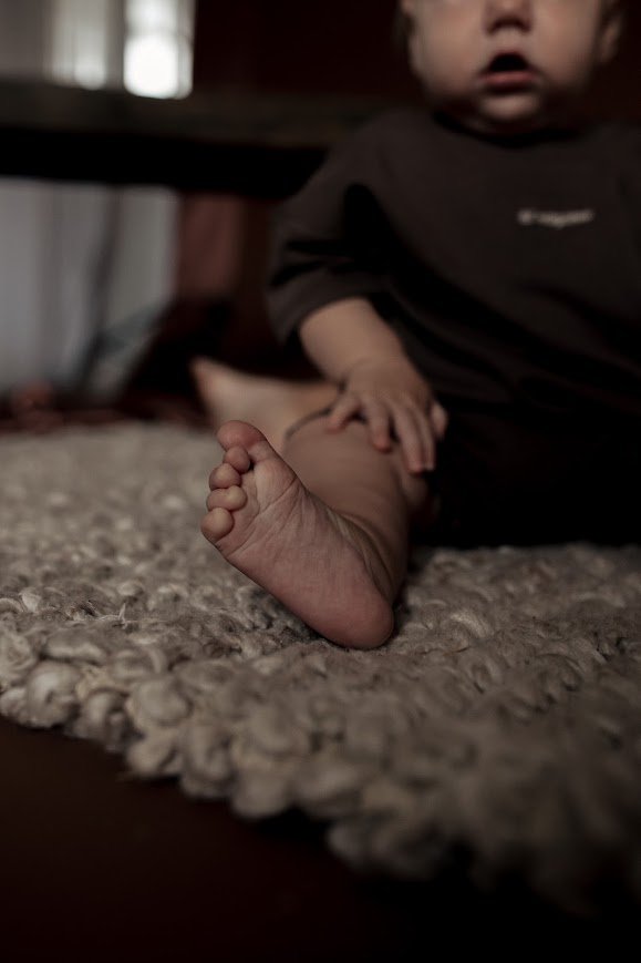 Close-up of a child's bare foot resting on a textured carpet while sitting on the floor, with part of the child's upper body visible in a dark shirt.