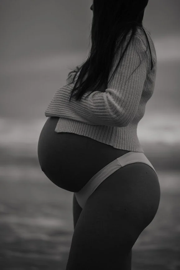 A pregnant woman in a gray sweater and underwear standing outdoors on the beach, facing sideways.