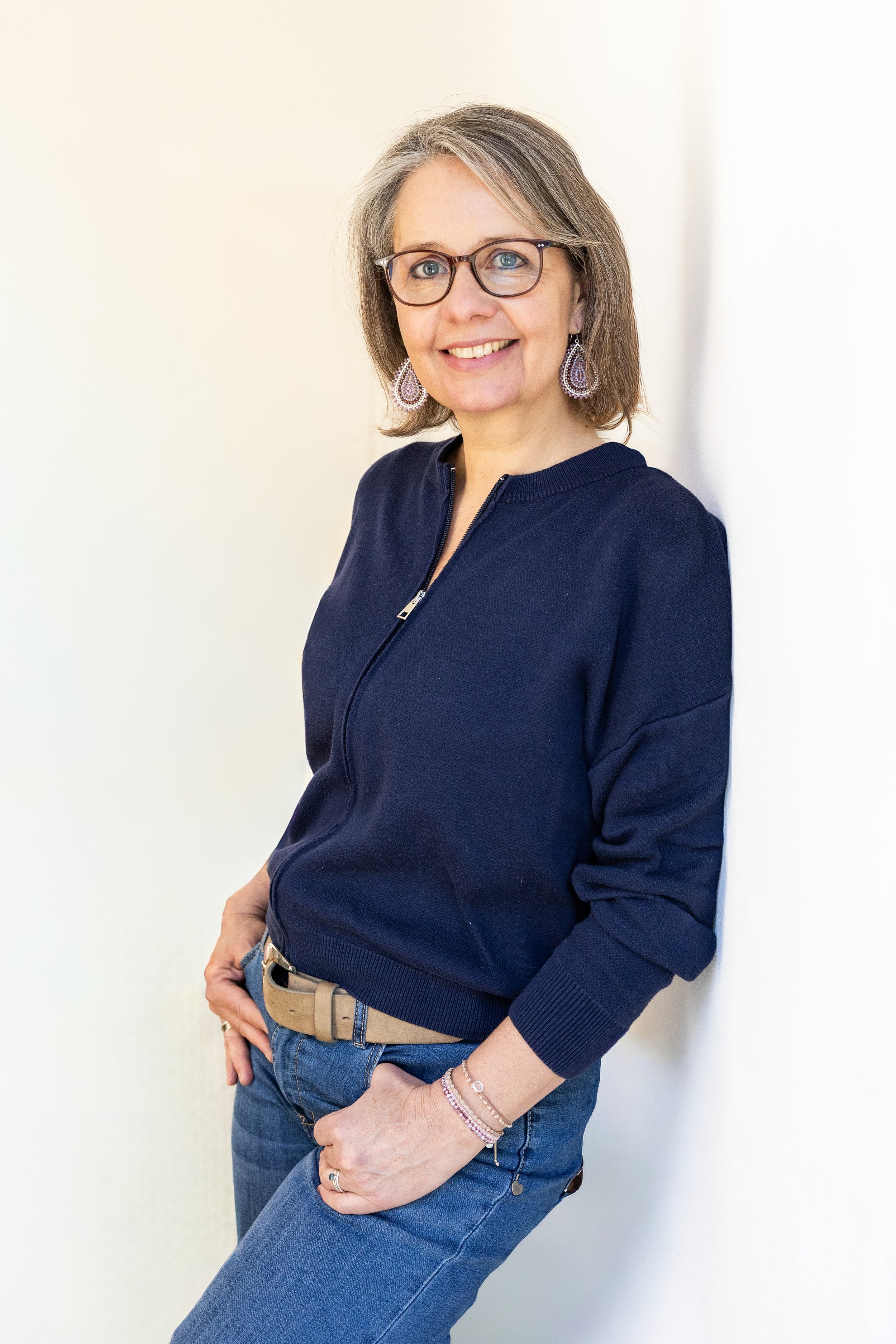 Eine lächelnde Frau mit Brille, kurzen grauen Haaren, trägt eine dunkelblaue Bluse, Jeans und Schmuck, steht vor einem hellen Hintergrund.
