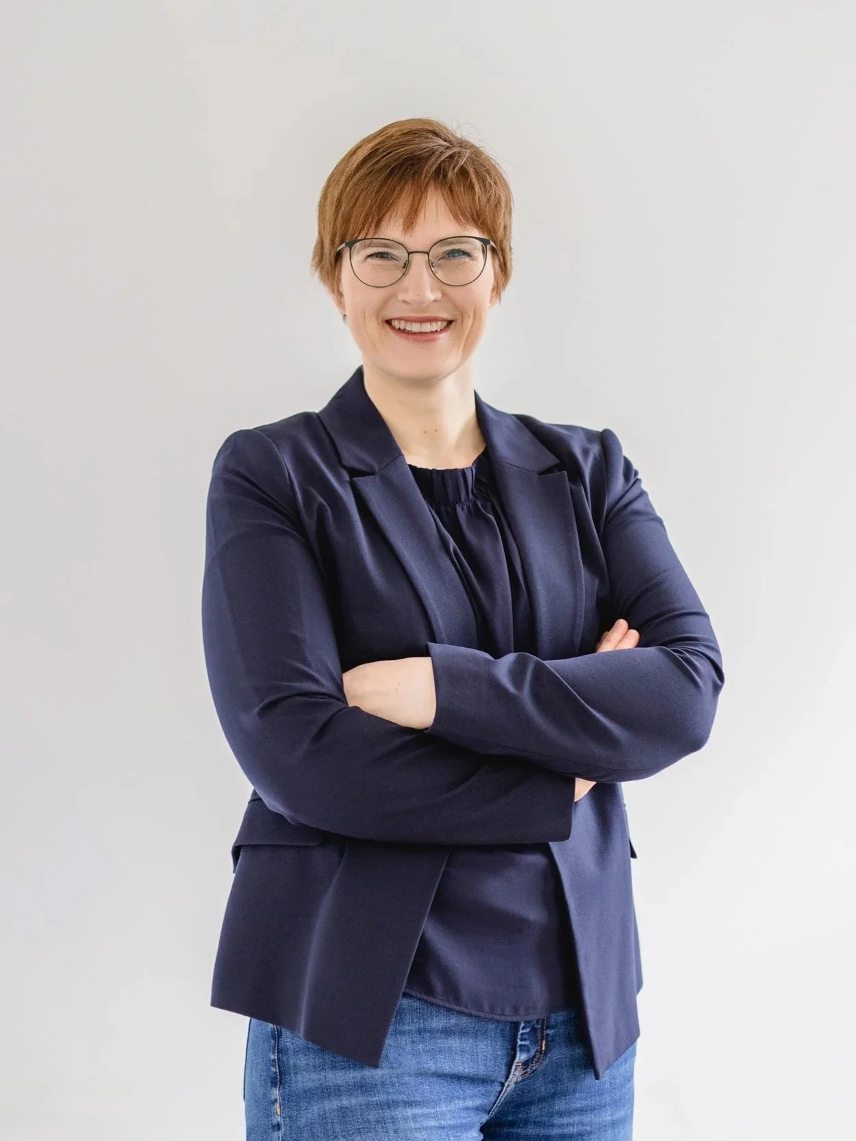 Eine Frau mit kurzen roten Haaren und Brille, trägt eine dunkelblaue Jacke und jeans, steht vor einer weißen Wand, lächelt und hat die Arme verschränkt.