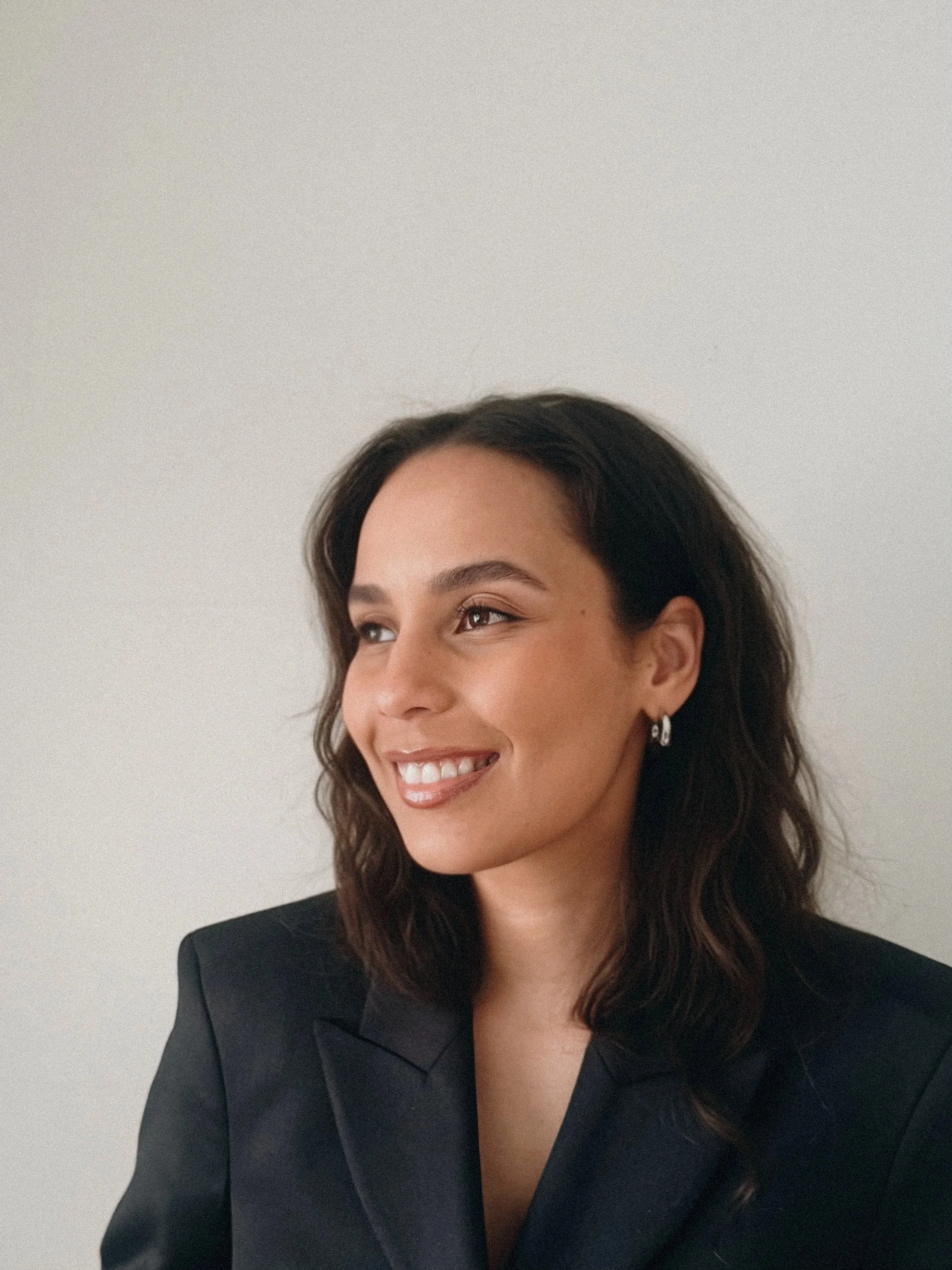A woman with dark hair smiling and looking to her left, wearing a black blazer and earrings, against a plain white background.
