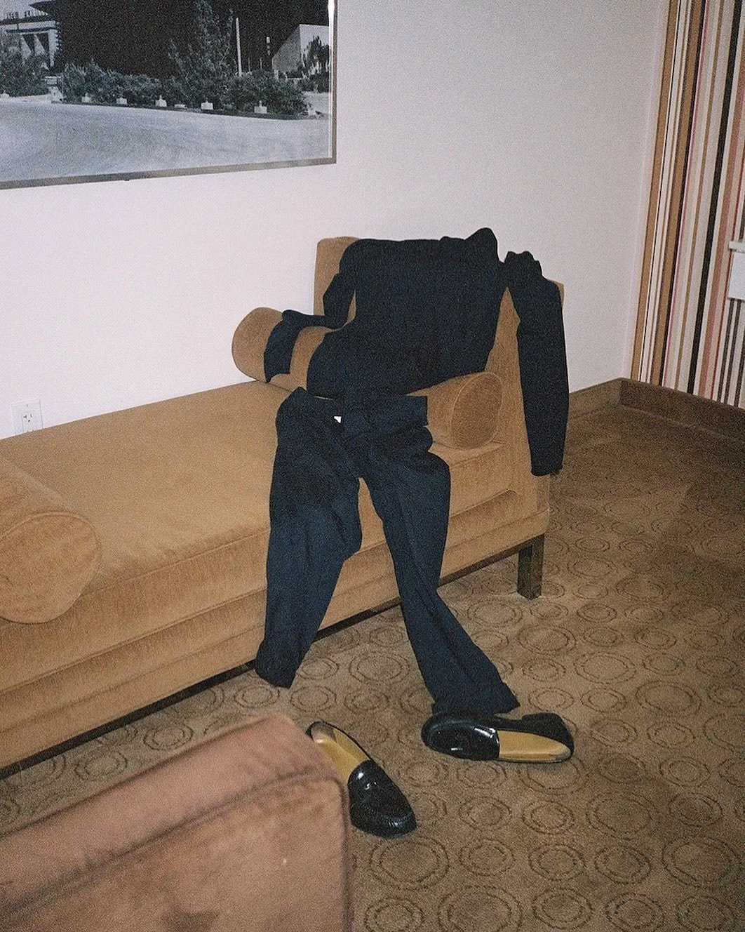 Clothes and shoes hanging on a beige sofa in a room with patterned carpeting and a large window.