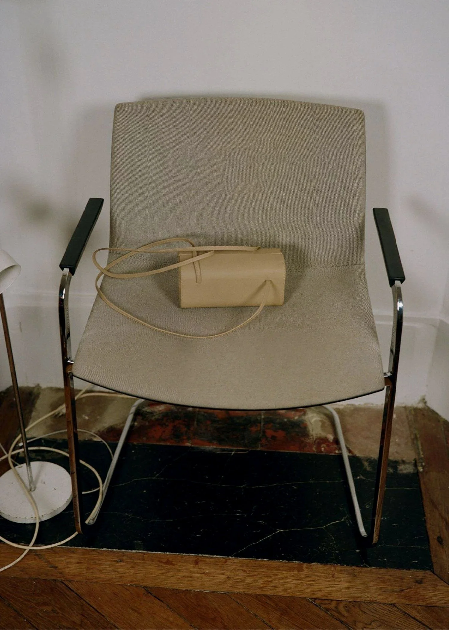 A beige chair with armrests, a small beige purse with a strap placed on it, and a floor lamp on the left side, all against a plain white wall with wooden flooring.