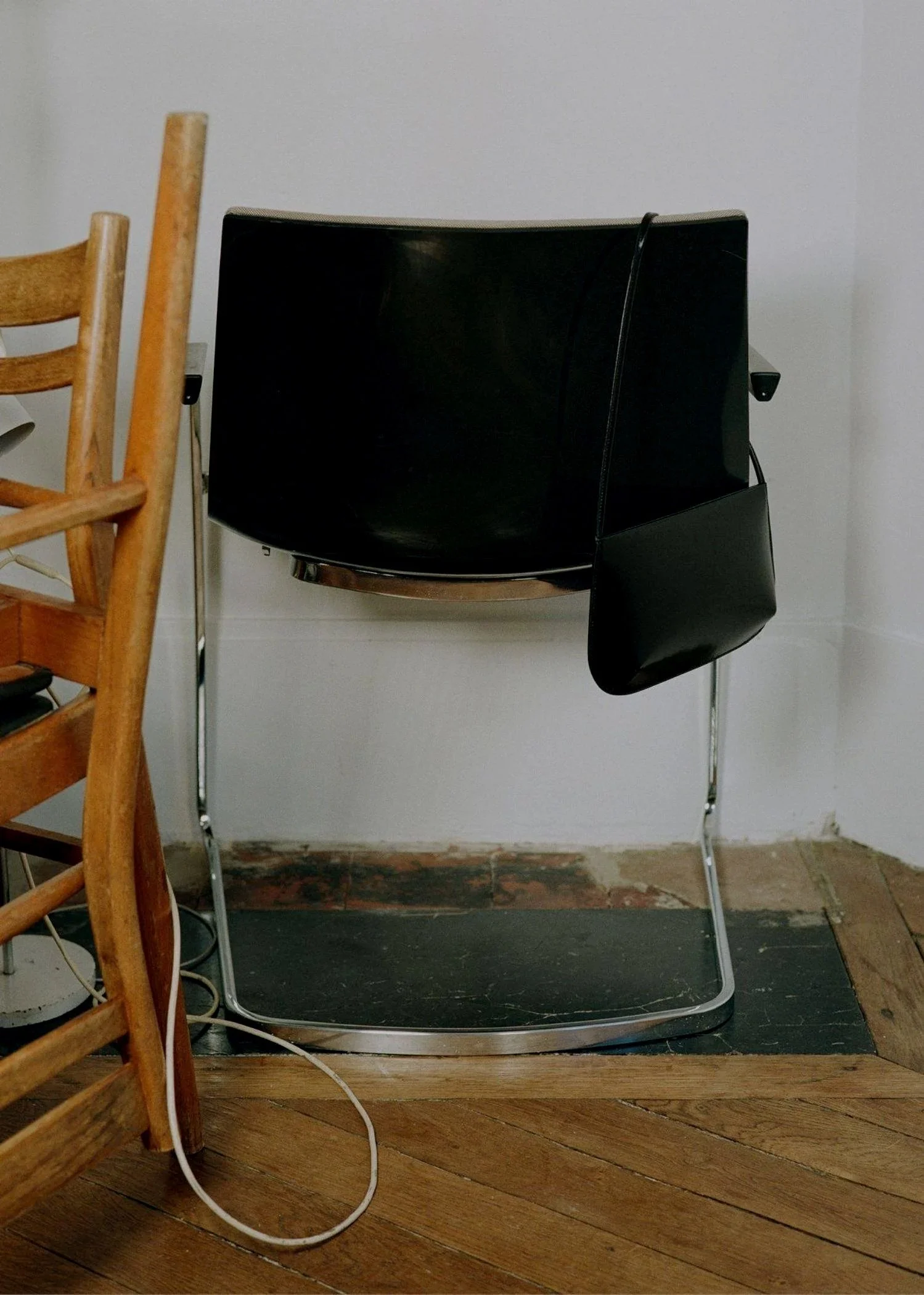Black chair with a small black bag hanging from it, situated against a white wall, on a black mat on wooden floor, with part of a wooden chair visible on the left.