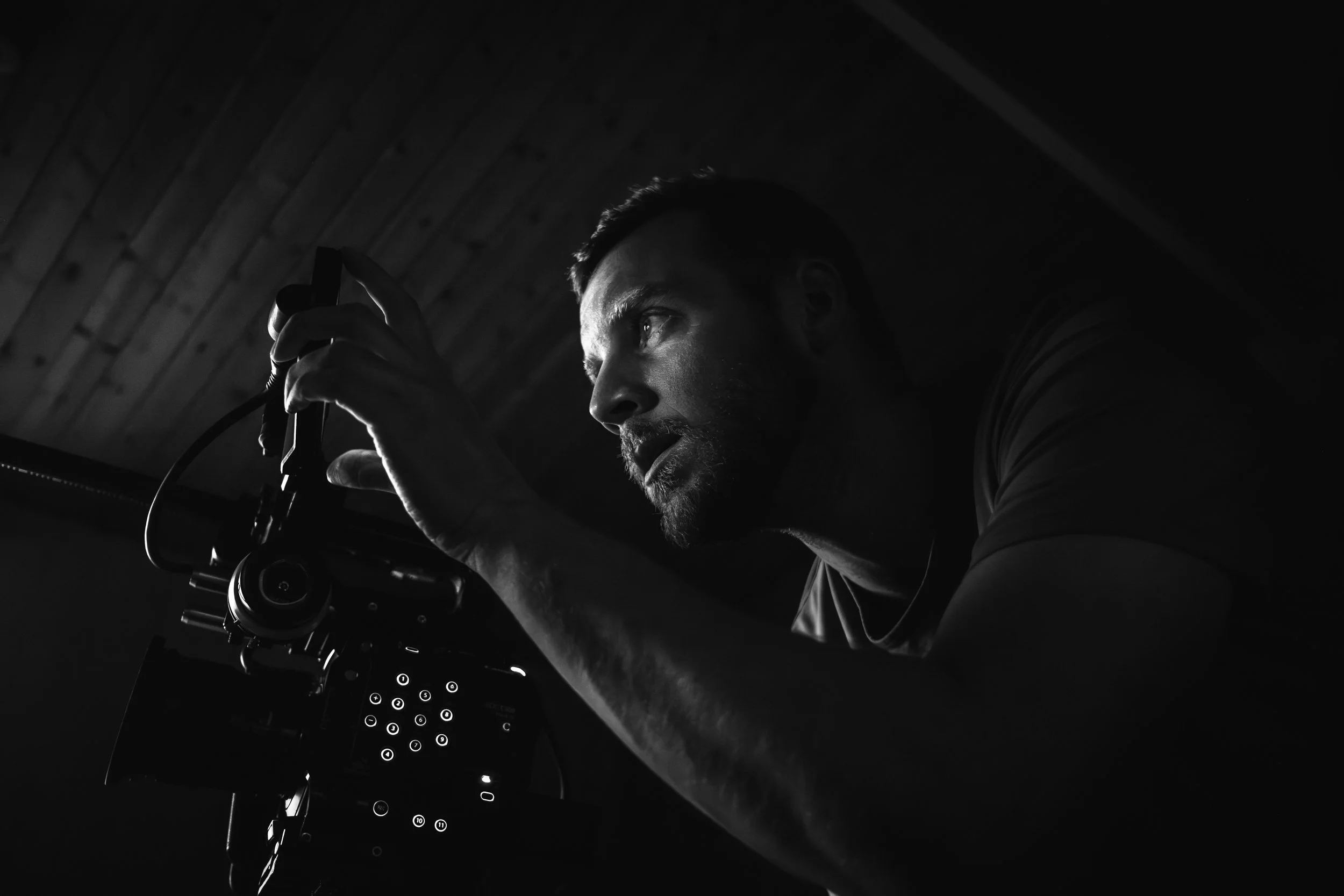 A man working with a camera or camera equipment in a dimly lit room, with a wooden ceiling in the background.