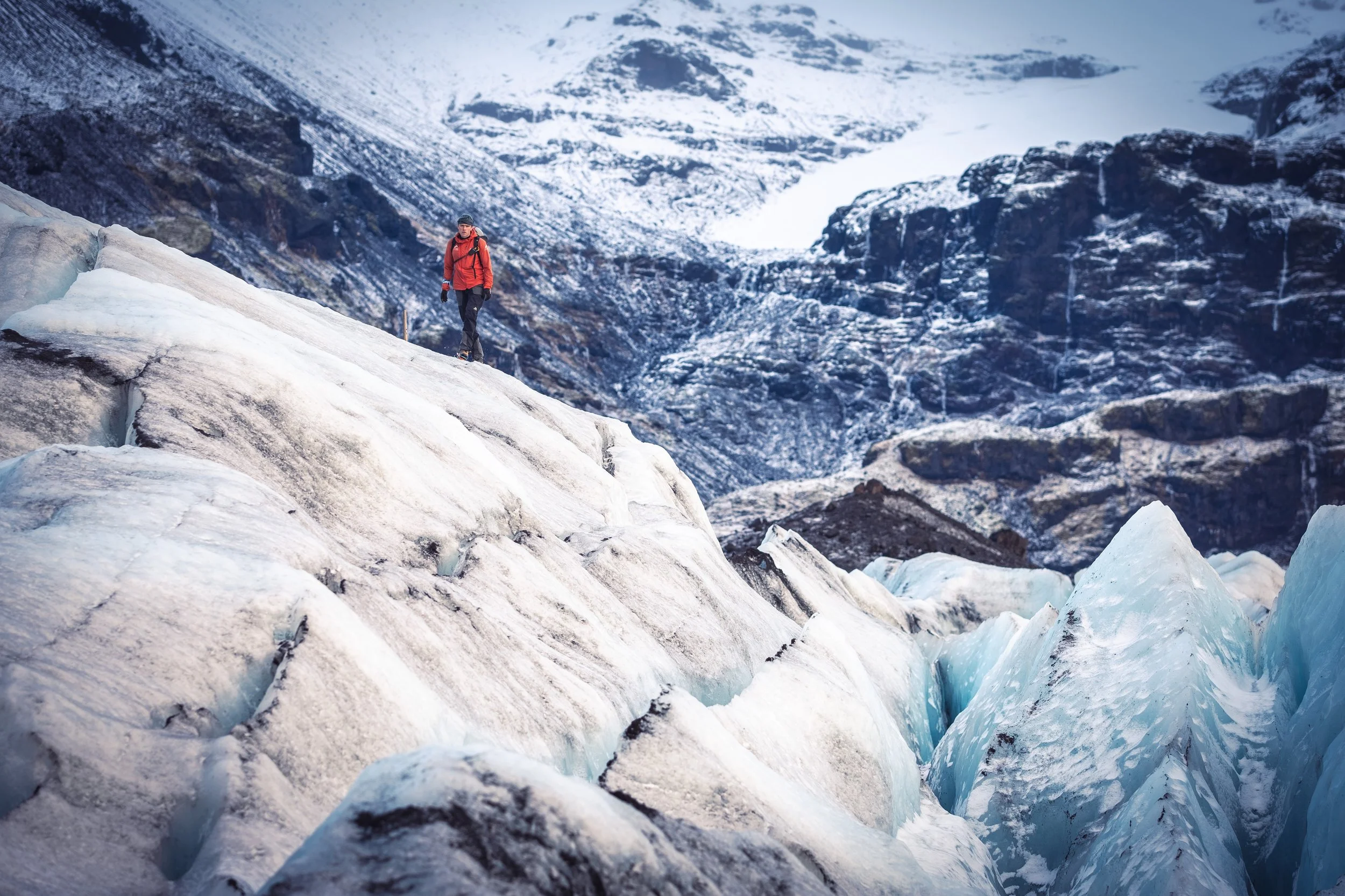 A person in a red jacket and black pants walking on icy terrain surrounded by snow-covered mountains.