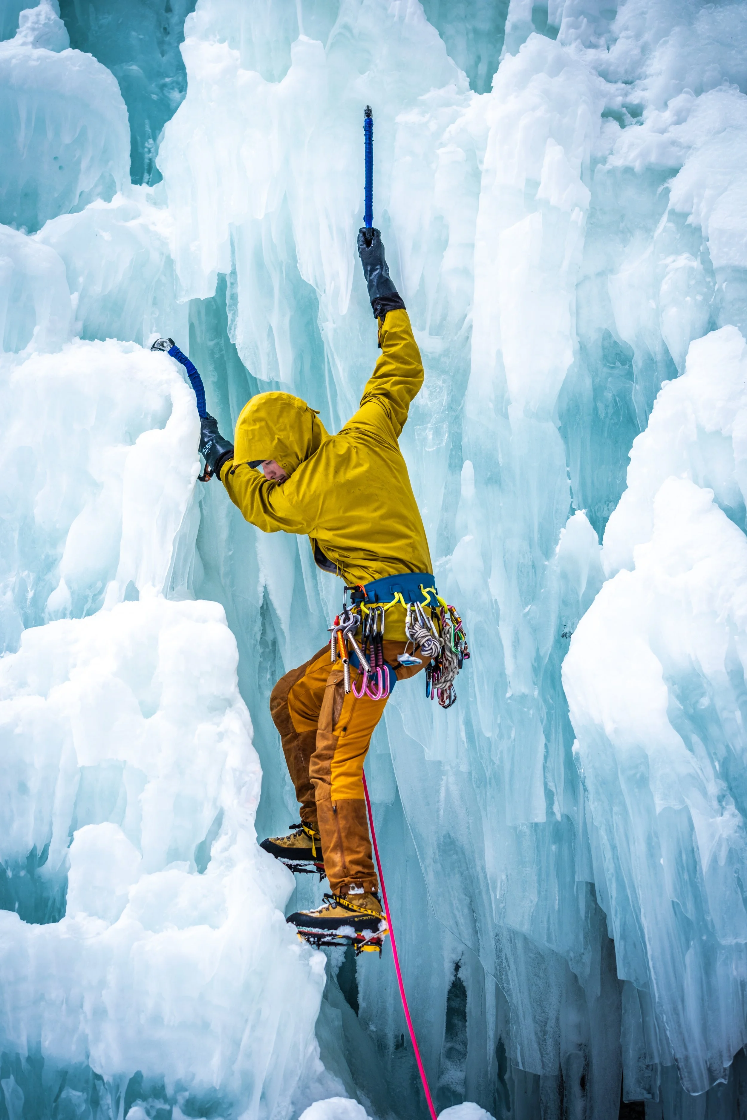 A person in a yellow jacket and brown pants, equipped with climbing gear, is ice climbing on a frozen glacier with ice axes and crampons.