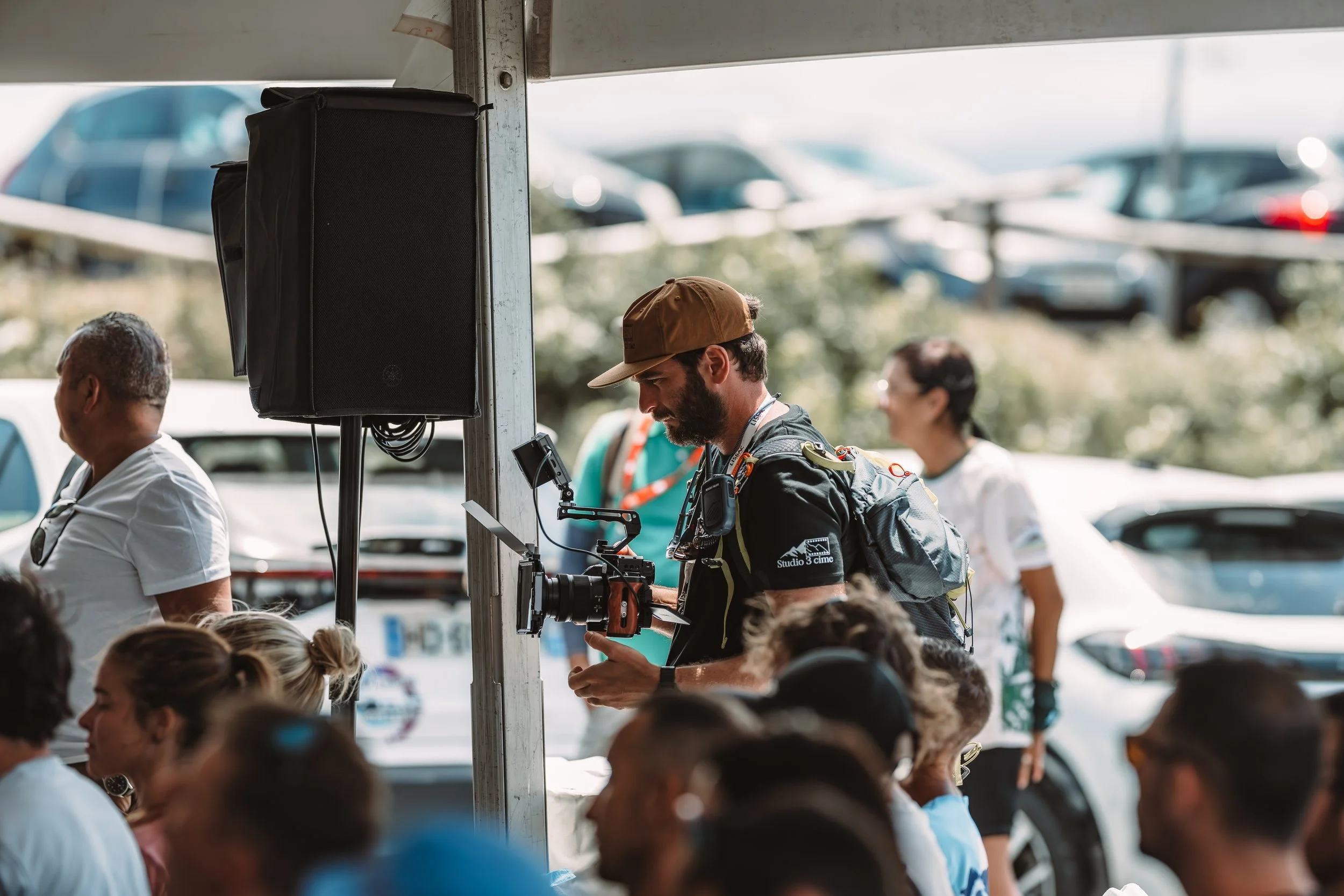 Cameraman with equipment recording at a gathering or event outdoors, with several seated and standing people, parked cars in the background.