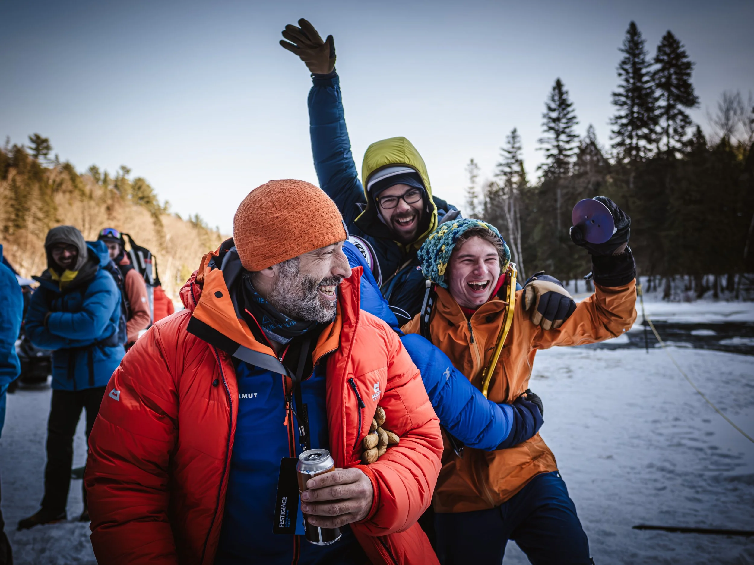 A group of people in winter outdoor clothing celebrating near a snowy river and trees, with joyful expressions.
