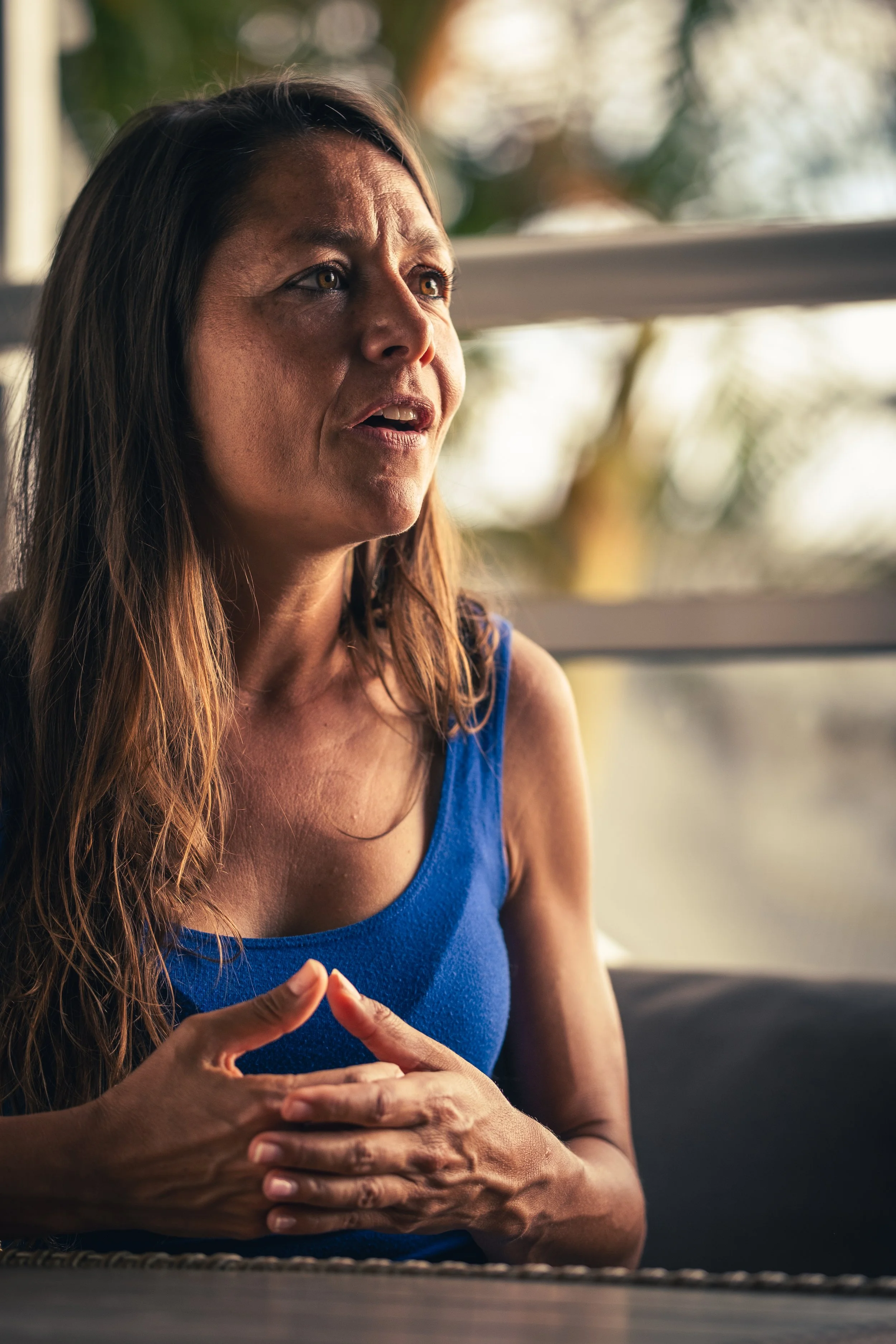 A middle-aged woman with long brown hair, wearing a blue sleeveless top, appears to be upset or worried, sitting indoors with natural light and greenery in the background.