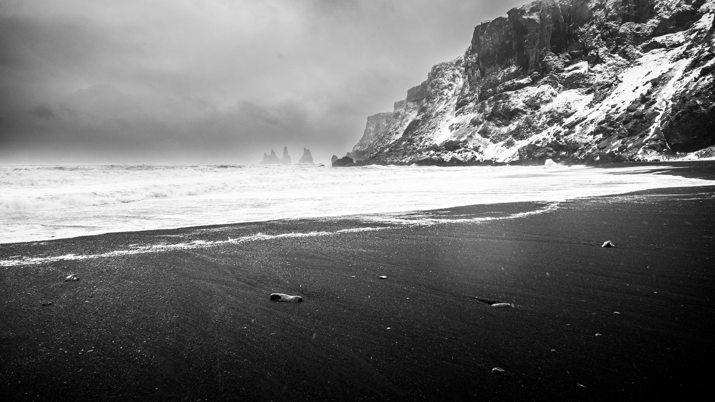 Black and white photograph of a rocky seaside with cliffs on the right, snowy and rugged. The shoreline is dark with scattered rocks, and waves are gently crashing onto the sand. The sky is overcast with a moody atmosphere.