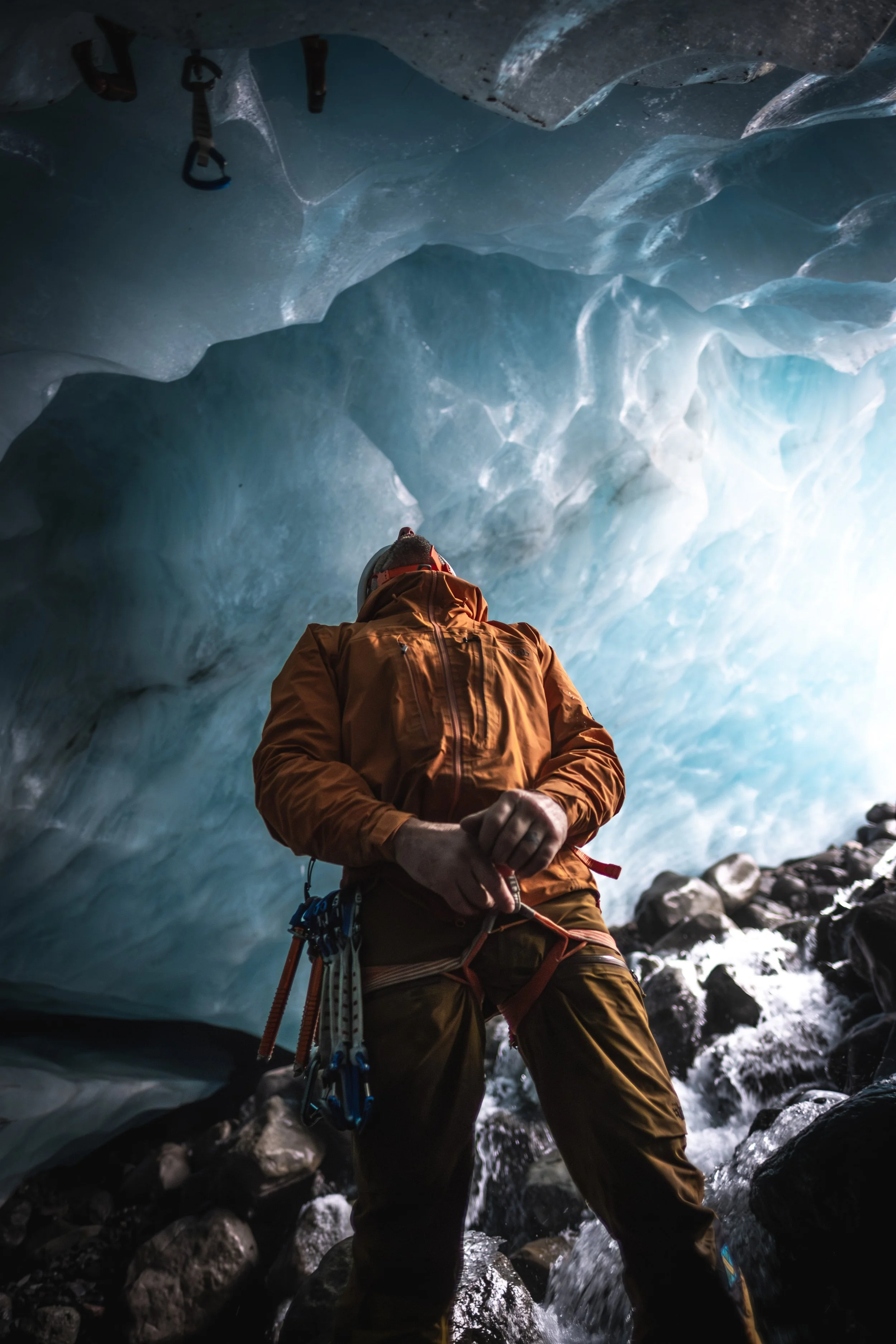 A mountain climber dressed in an orange jacket and gear standing inside an ice cave with icy blue walls and rocks on the ground.