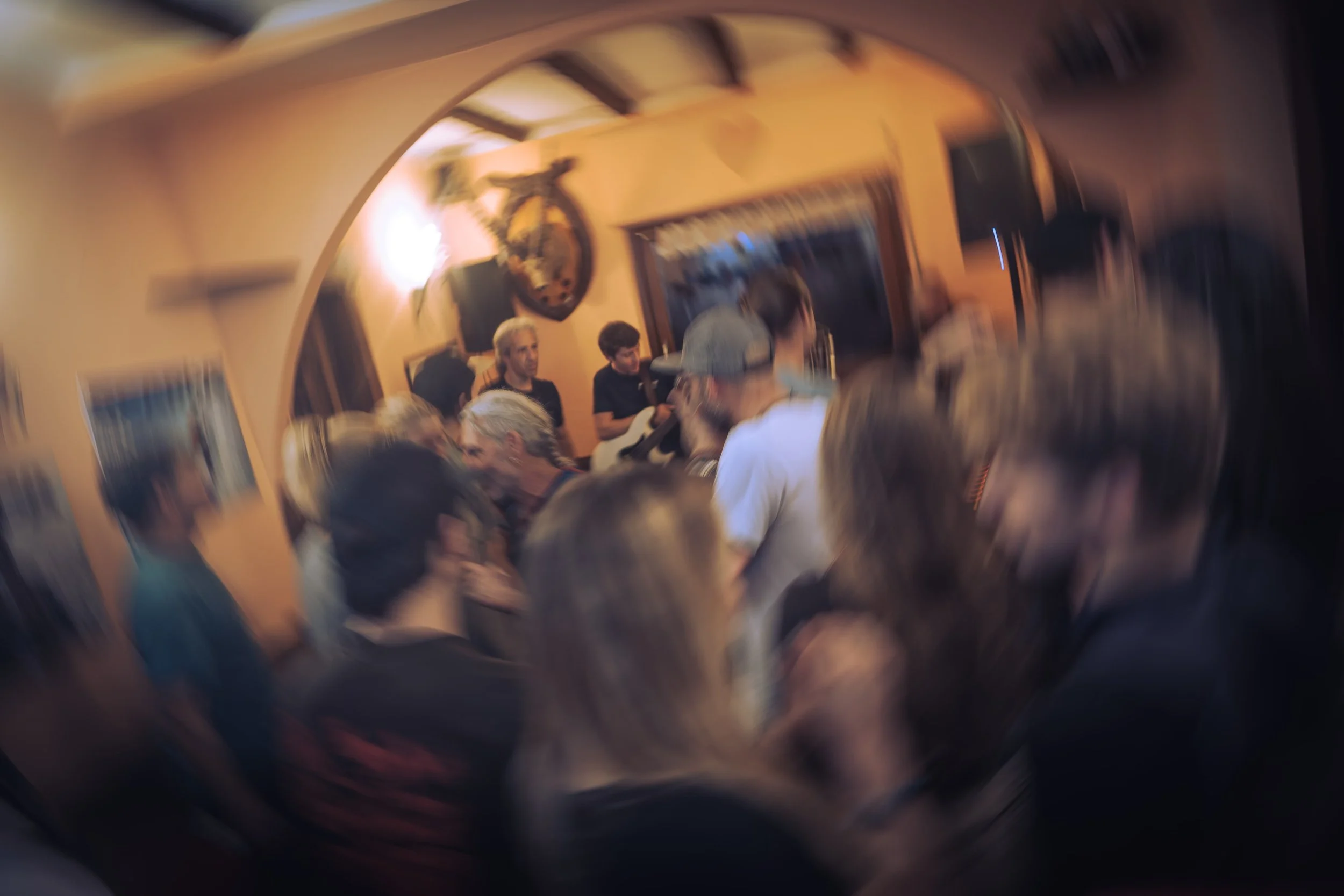 A crowded indoor gathering with a band performing in the background, motorcycles on the wall, and various people talking and socializing.