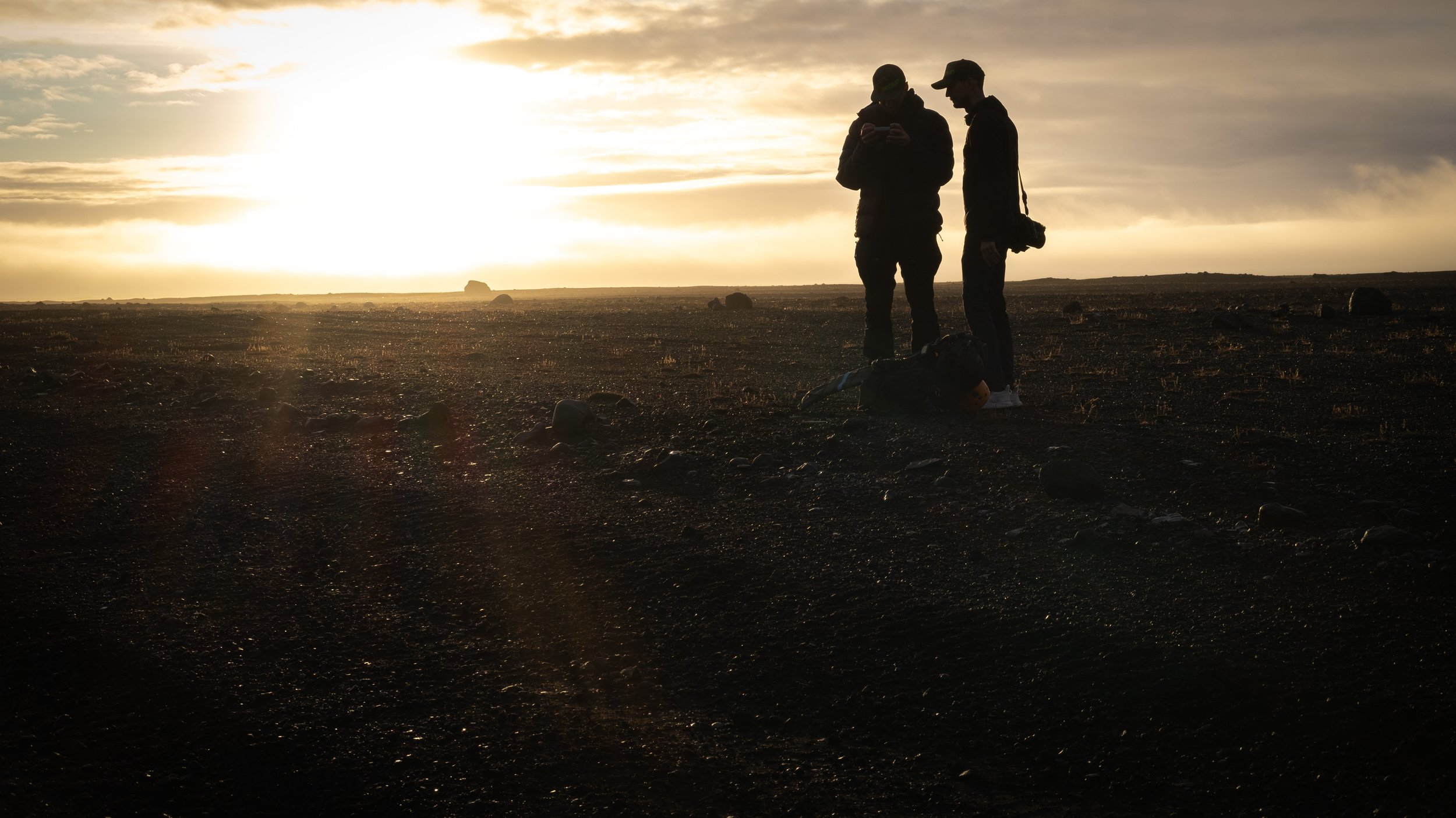 Two people standing outdoors at sunset, silhouetted against the sky, with a camera on the ground nearby.