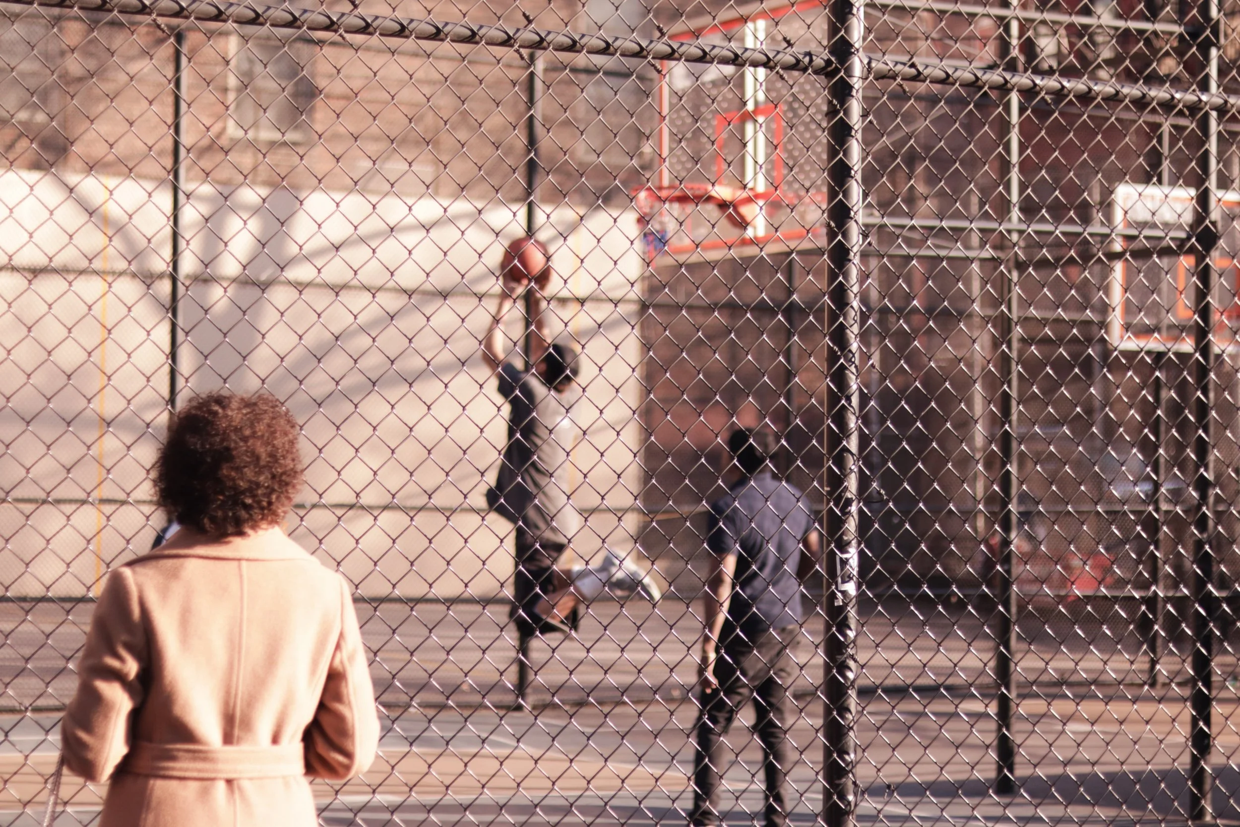 A basketball court behind a chain-link fence with two people playing basketball and a woman watching from outside the fence.