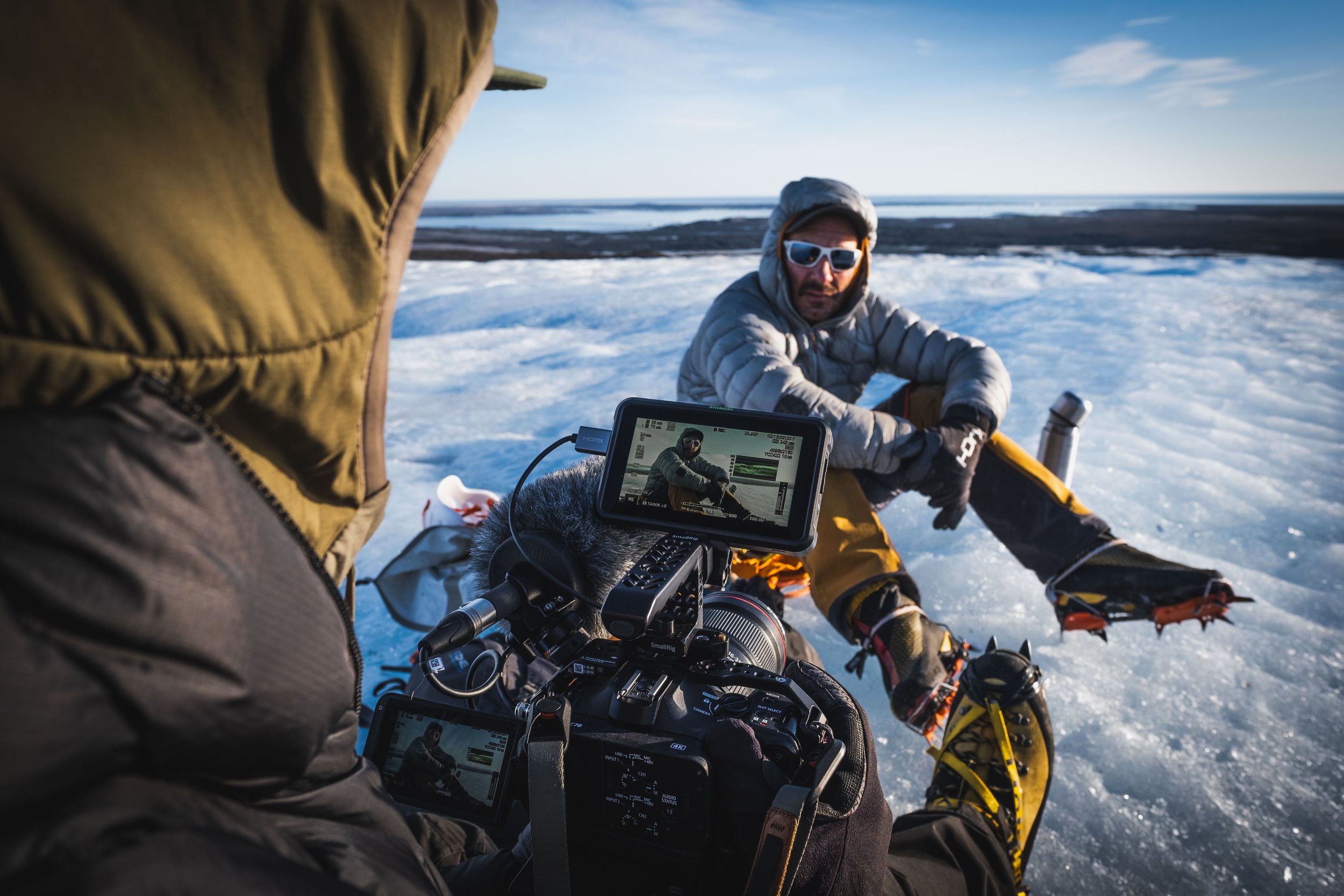 Two explorers in winter gear on a snowy icy landscape, with one person filming the other using a professional camera setup.