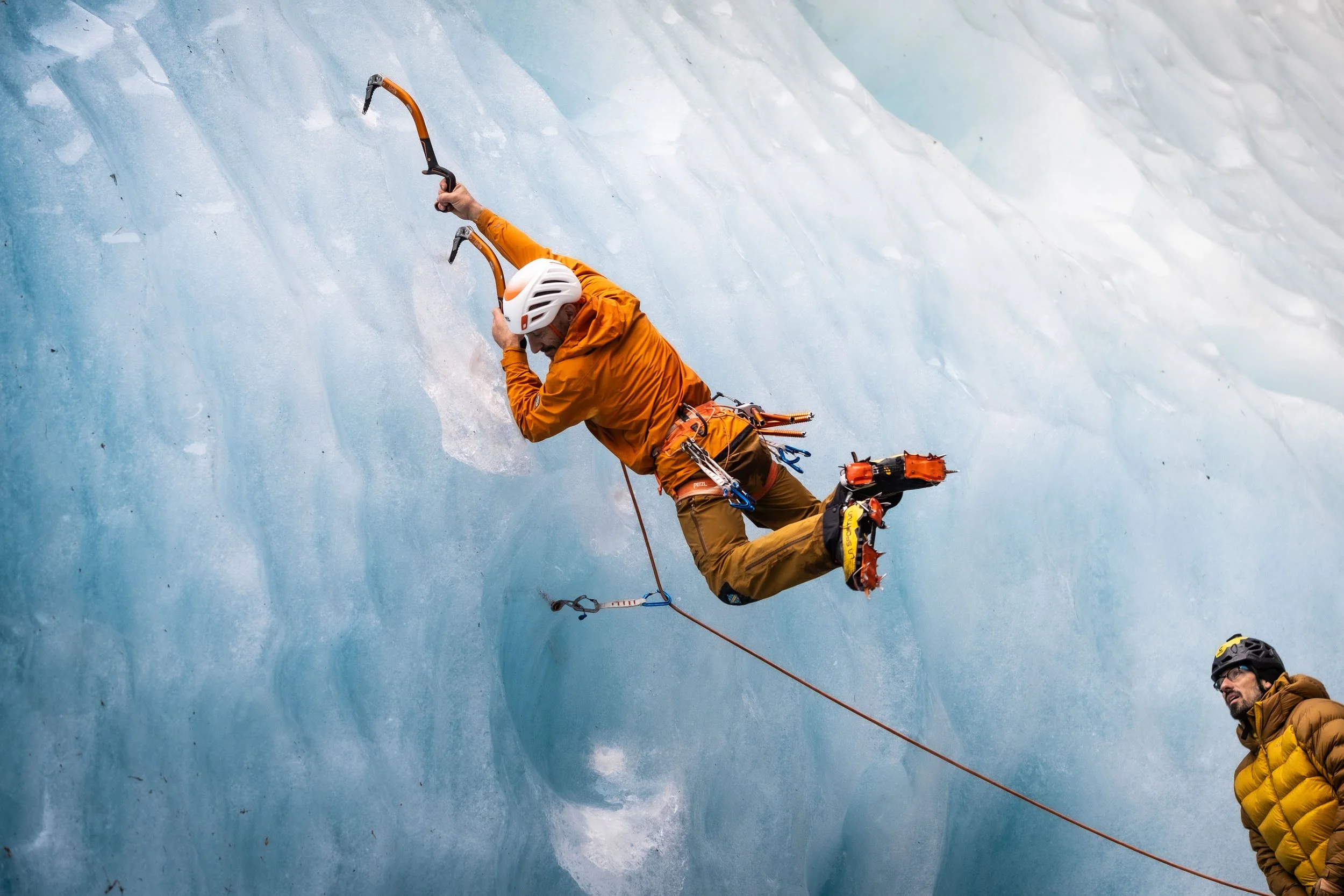 Ice climber in orange jacket and helmet ascending icy wall with two other climbers nearby.