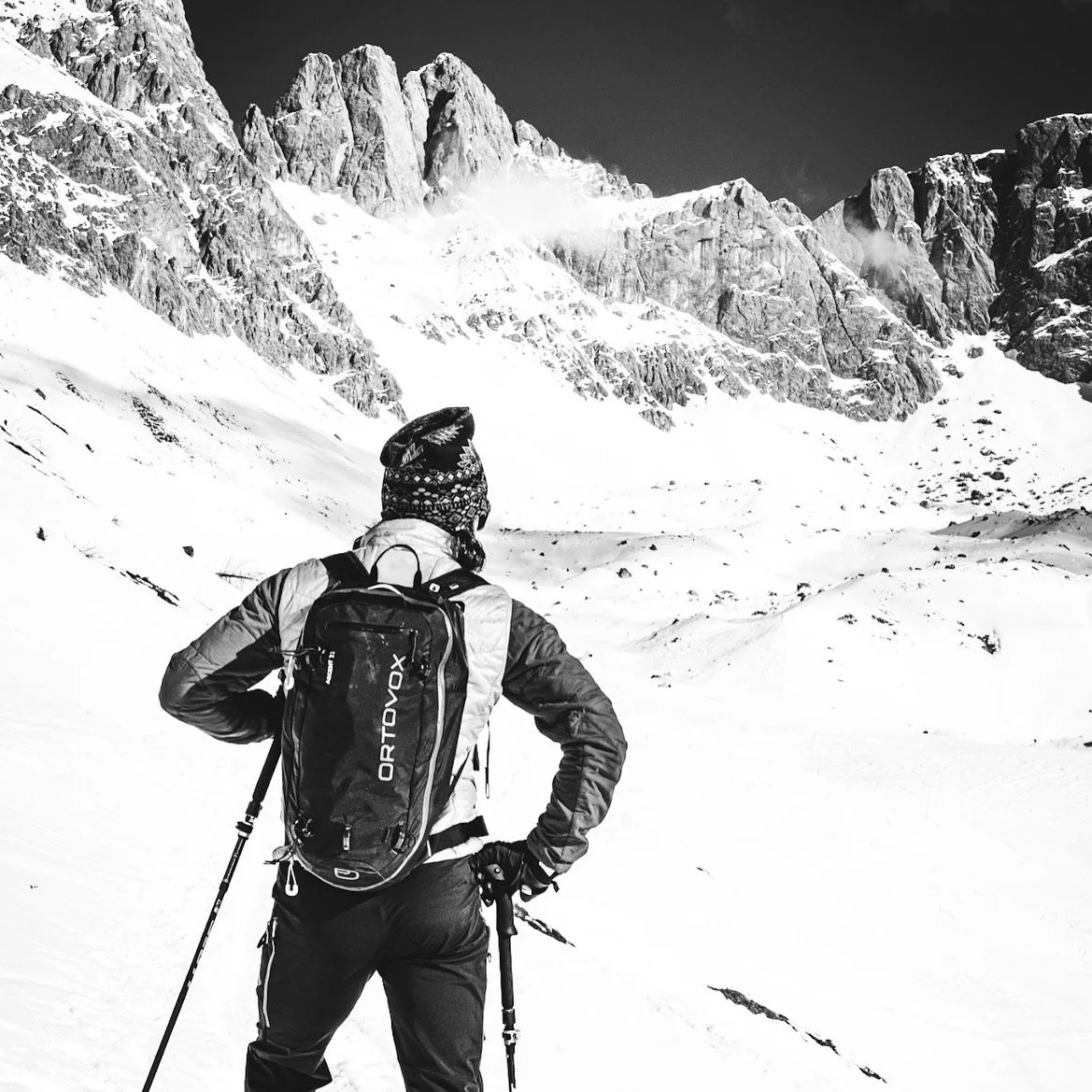 A person wearing winter gear, including a backpack and holding trekking poles, standing in a snowy mountain landscape with rugged, snow-covered peaks in the background.