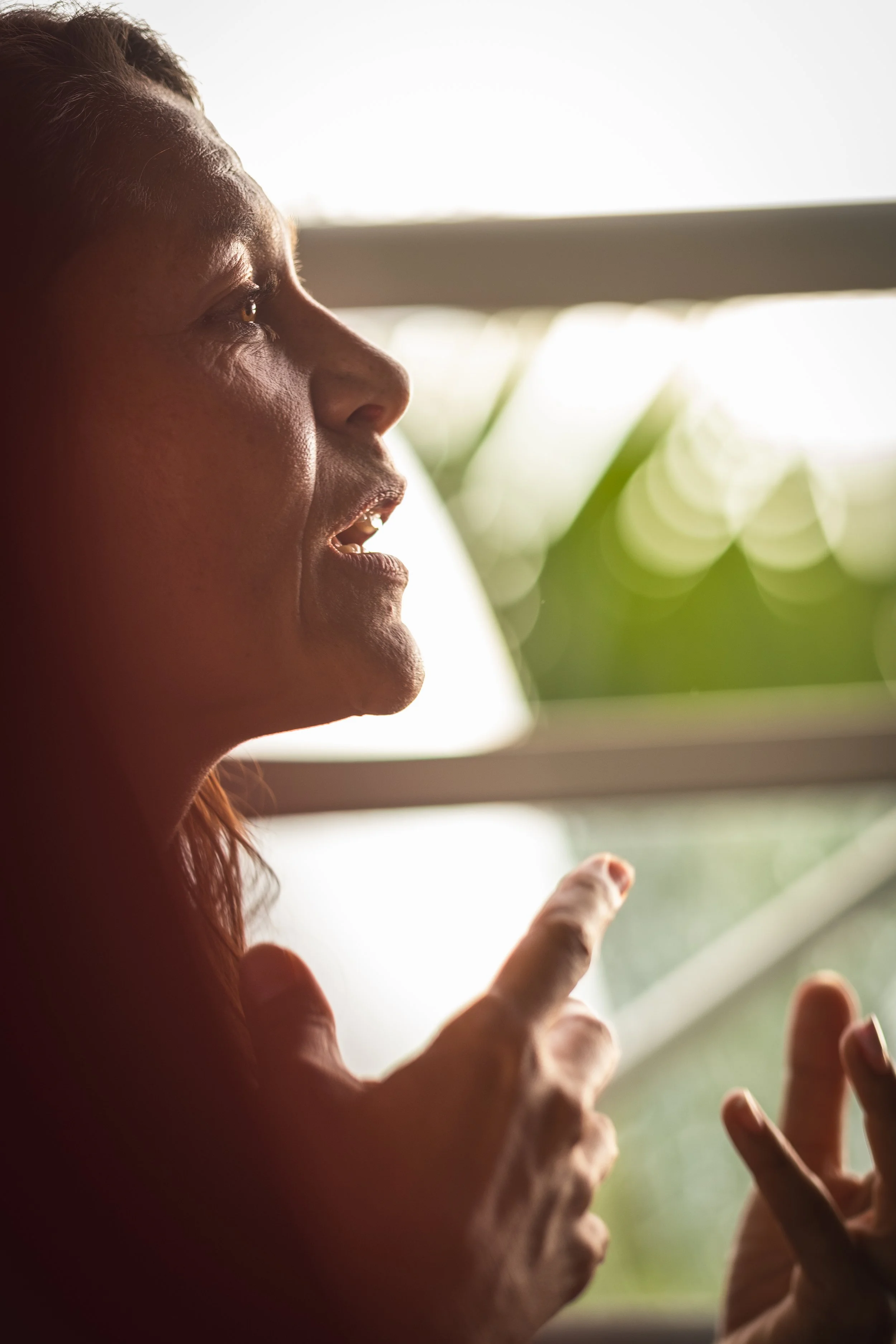 A woman with reddish hair and tan skin is talking near a window, with natural light illuminating her face and hands.