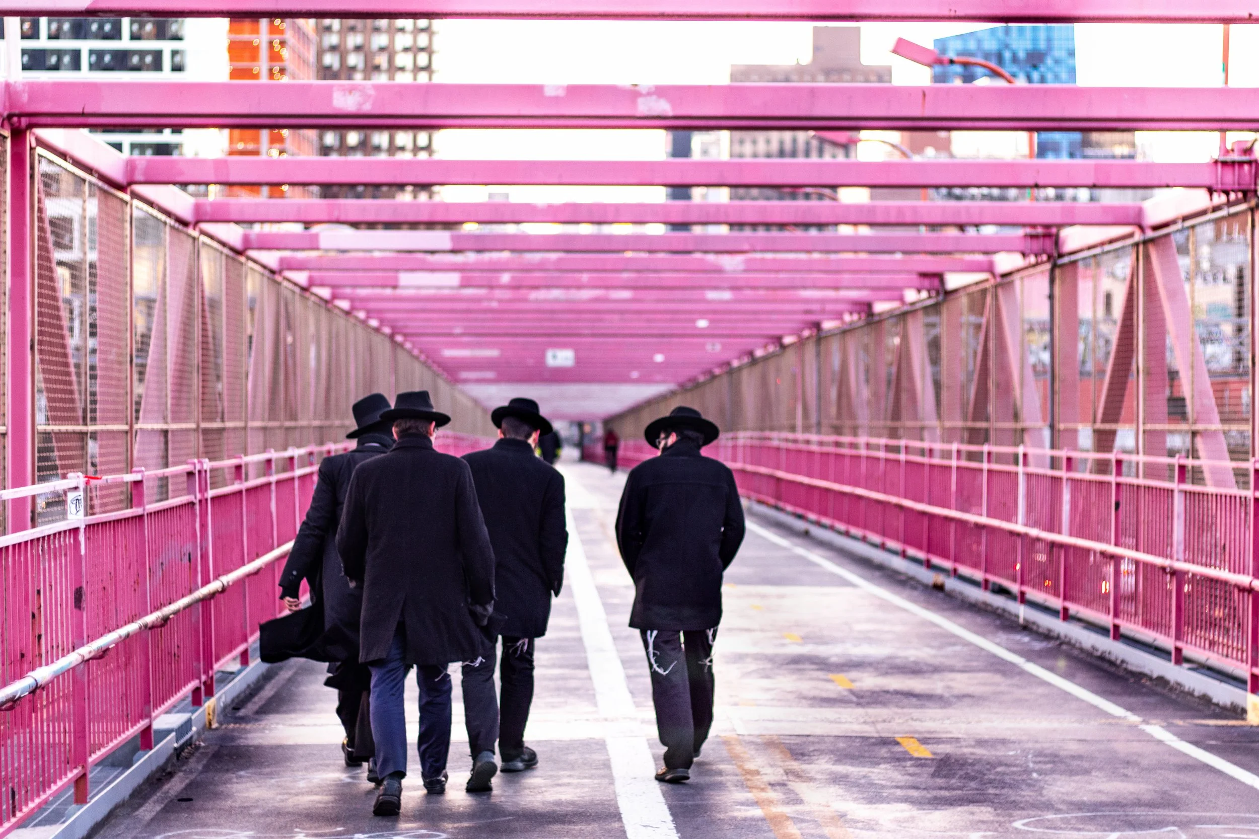 Four men in black coats and hats walking on a pink bridge in an urban area with high-rise buildings in the background.