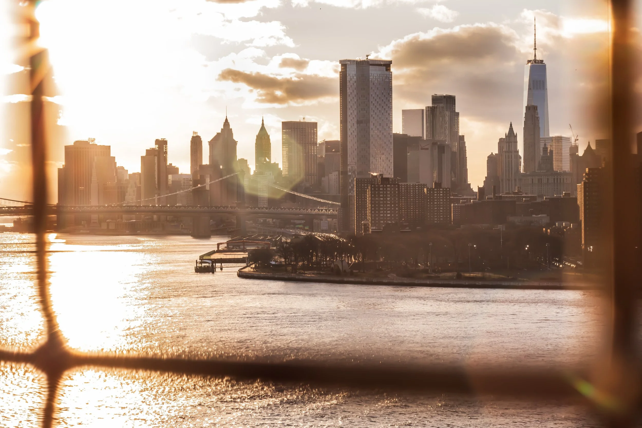 View of New York City skyline with tall skyscrapers, including One World Trade Center, seen from across a river during sunset.