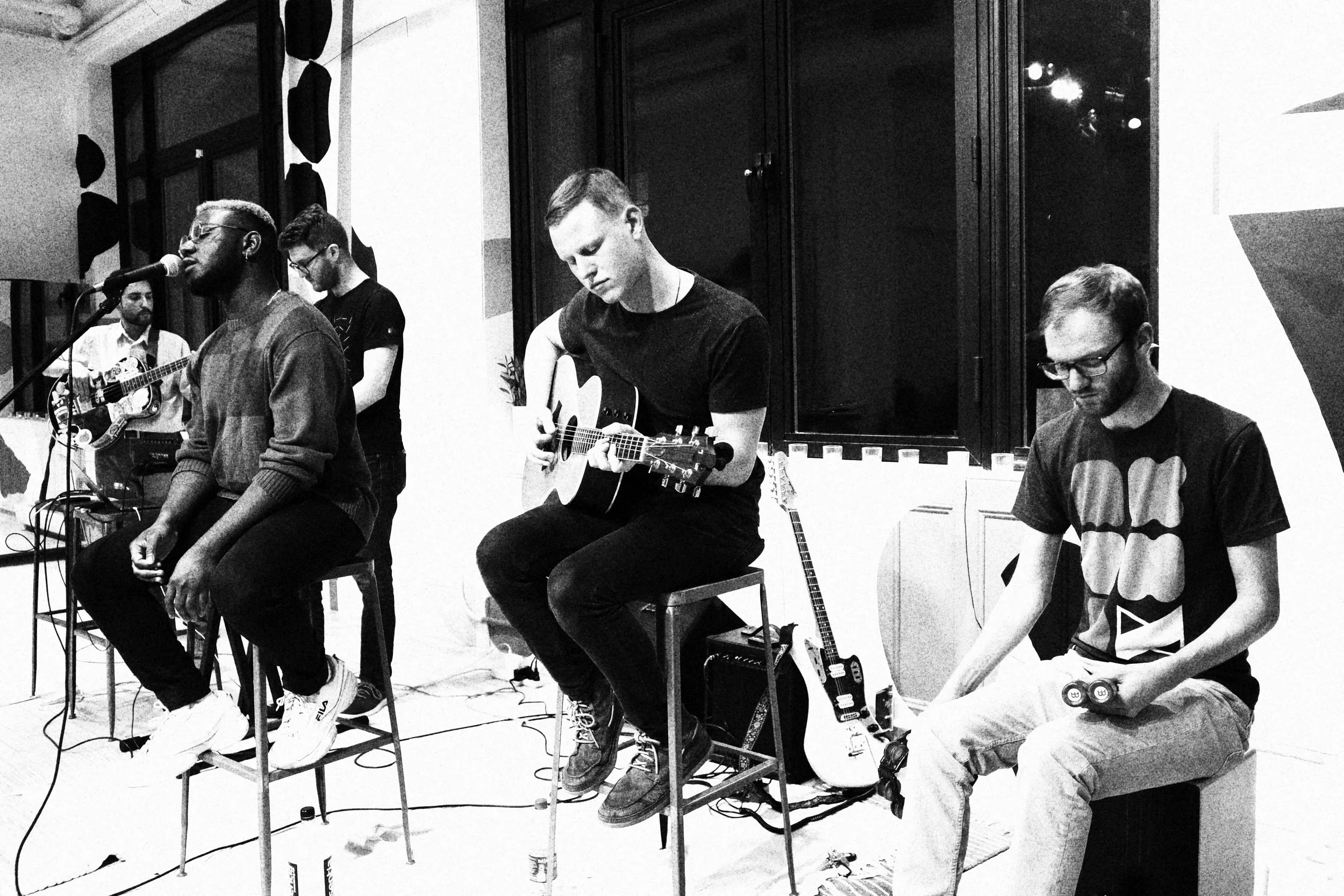 A group of five musicians performing indoors. One person is singing into a microphone, two are playing acoustic guitars, one is playing a cajón, and the last person's face is obscured.