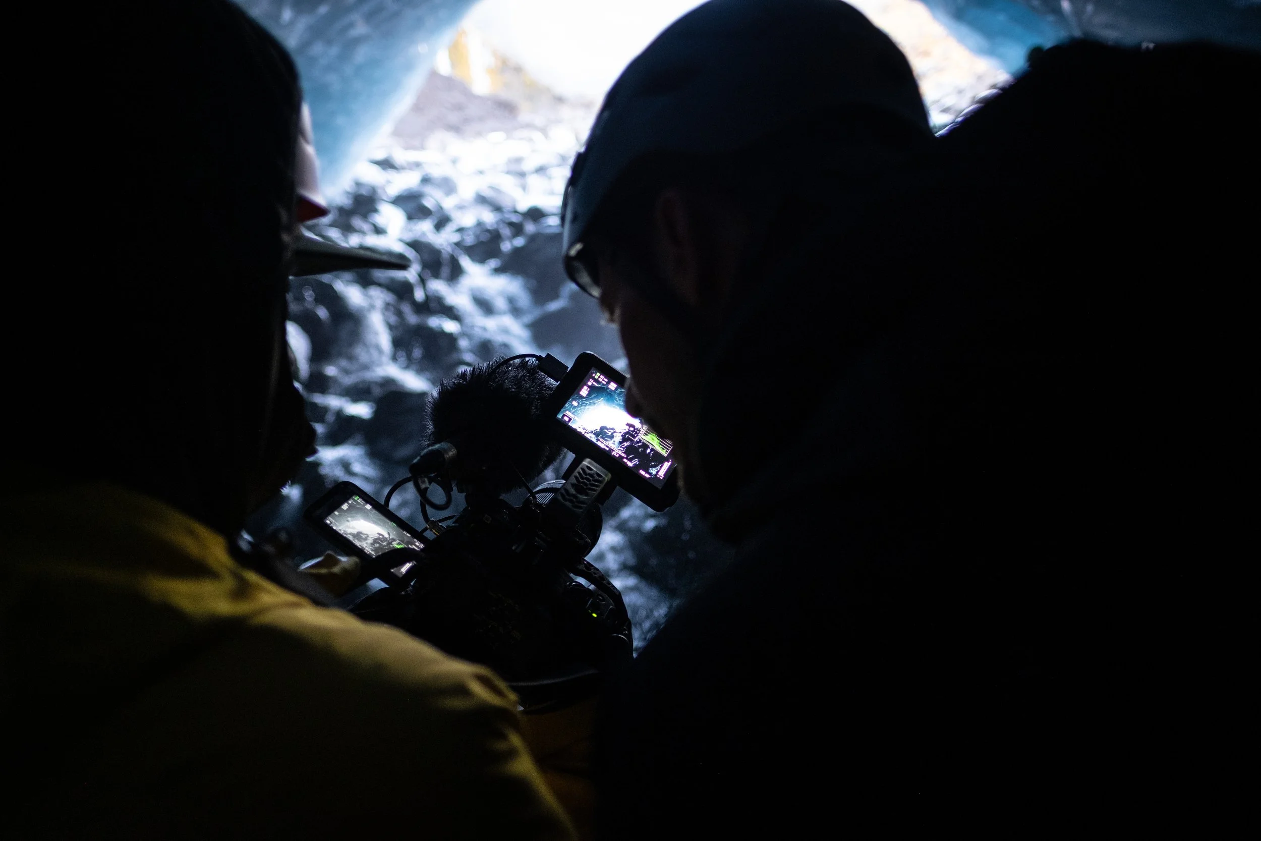 Two people in a dark cave with a stream of water, filming or taking photos of the cave walls.