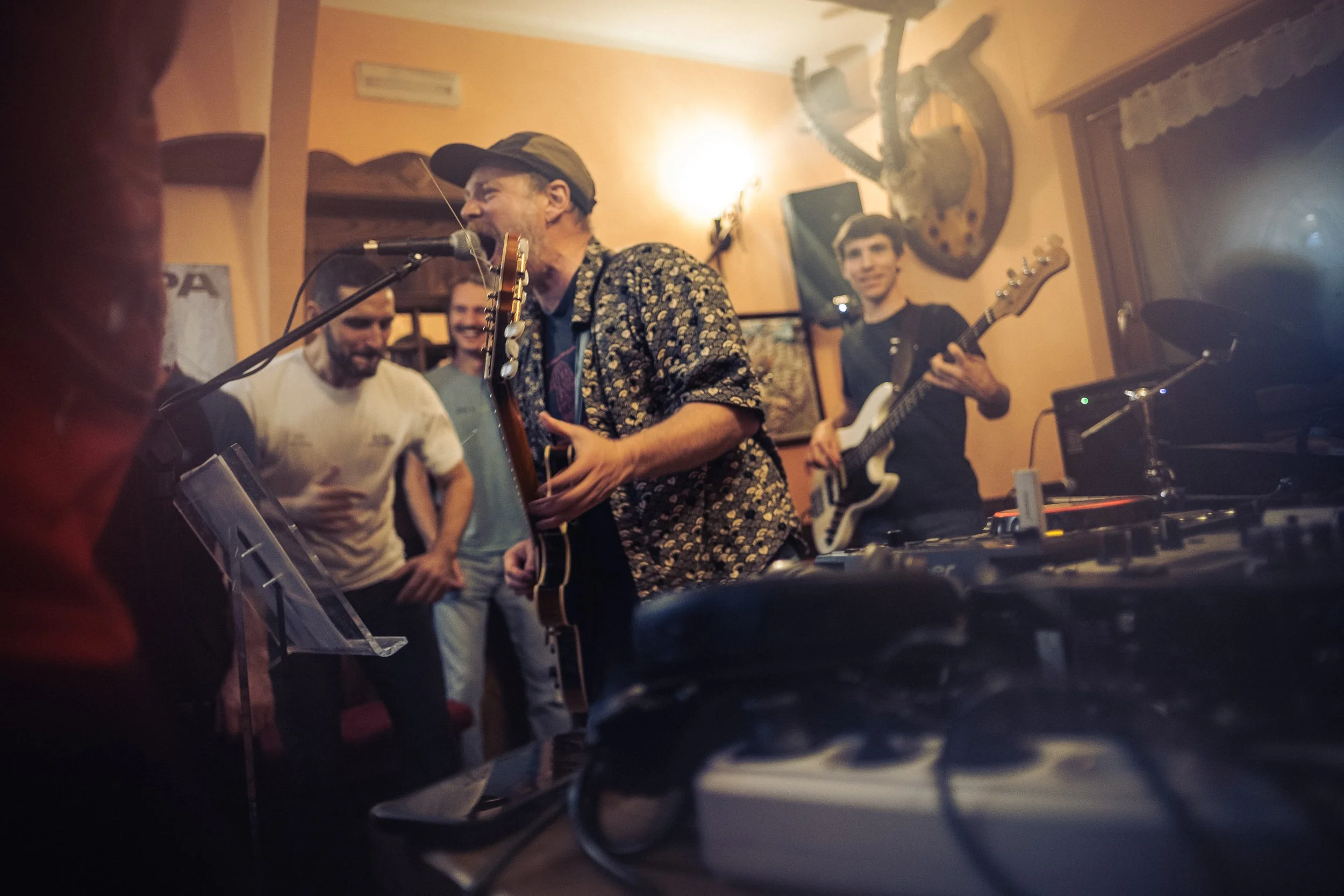 A band performing in a cozy indoor setting with warm lighting, featuring a singer with a guitar, an electric guitarist, and a few audience members enjoying the music.