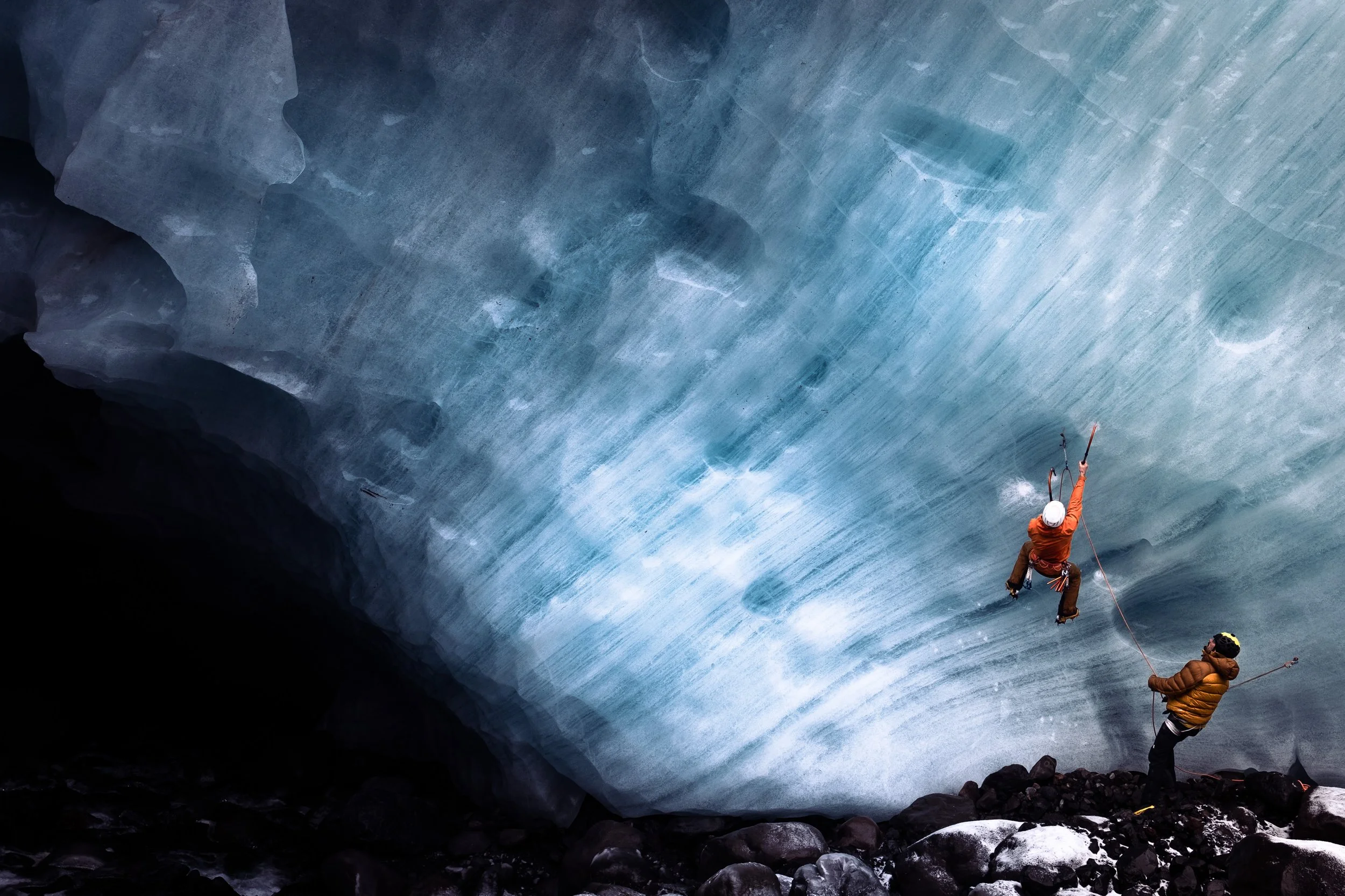 Two ice climbers on a glacier, one person wearing an orange jacket and helmet, the other in a brown jacket and helmet, using ropes and ice axes to ascend a blue ice wall.