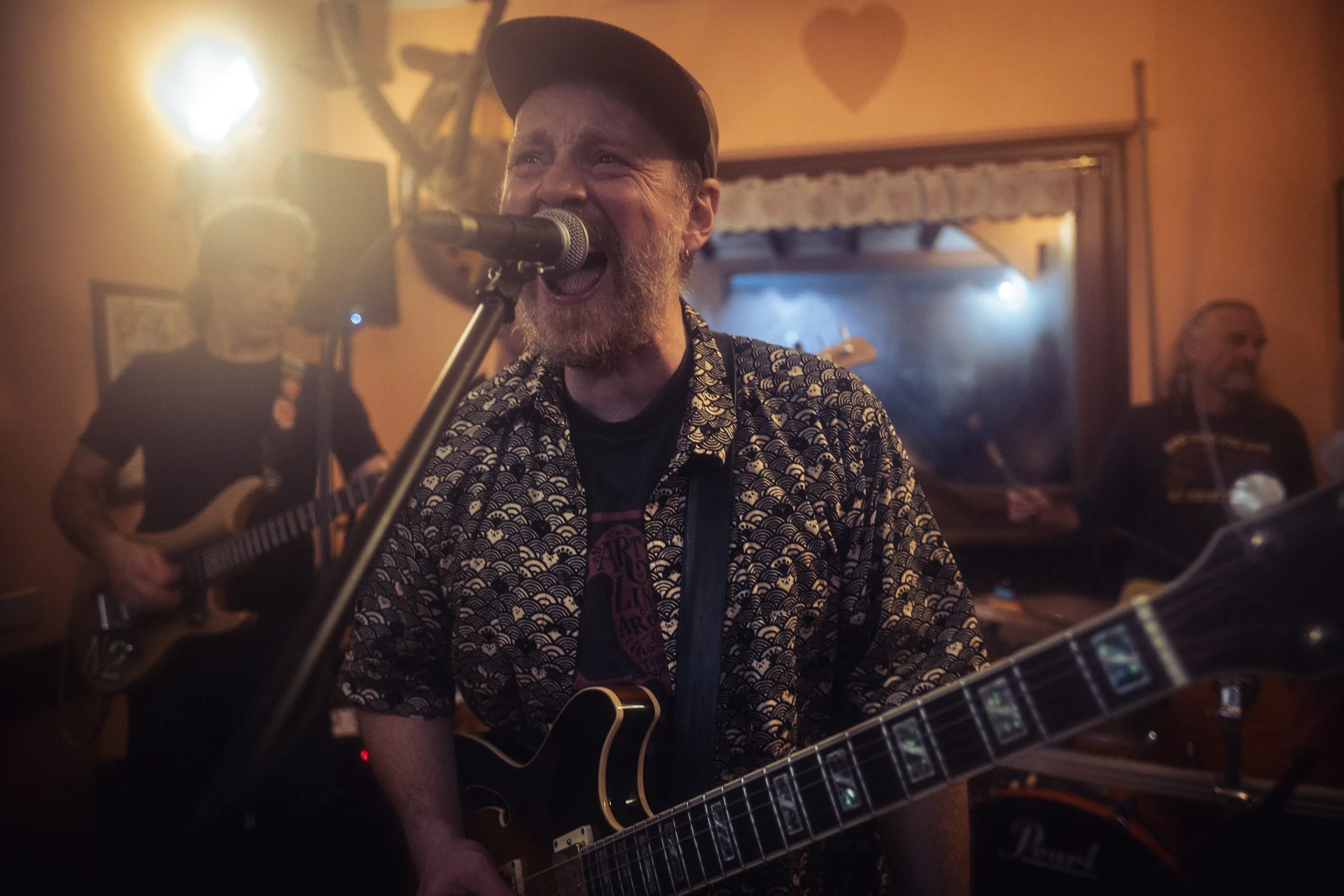 Musician passionately singing into a microphone while playing an electric guitar in a dimly lit room, with two other band members playing guitars in the background.