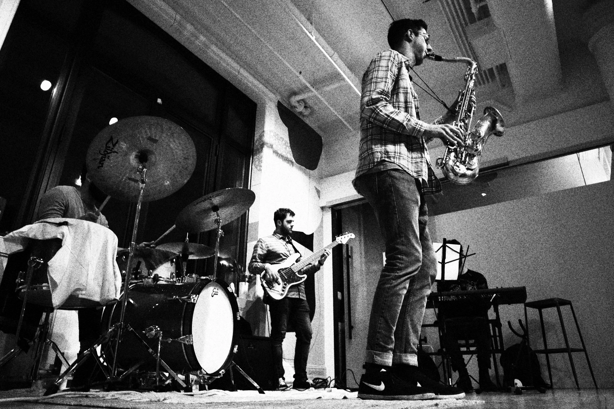 A black-and-white photo of a band performing indoors. There is a drummer on the left, a bass guitarist in the middle, and a saxophonist on the right. The band members are playing their instruments, with the saxophonist standing and the others sitting