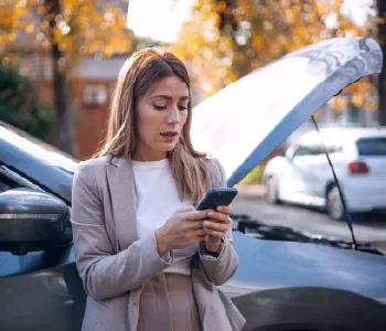 Woman standing outdoors, looking at her phone with an open car trunk in the background.