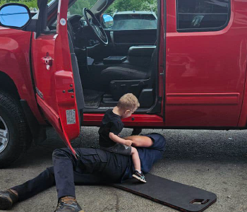 Child sitting on an adult lying on the ground next to an open car door, with the adult underneath the vehicle. The car is red, parked on a paved surface.