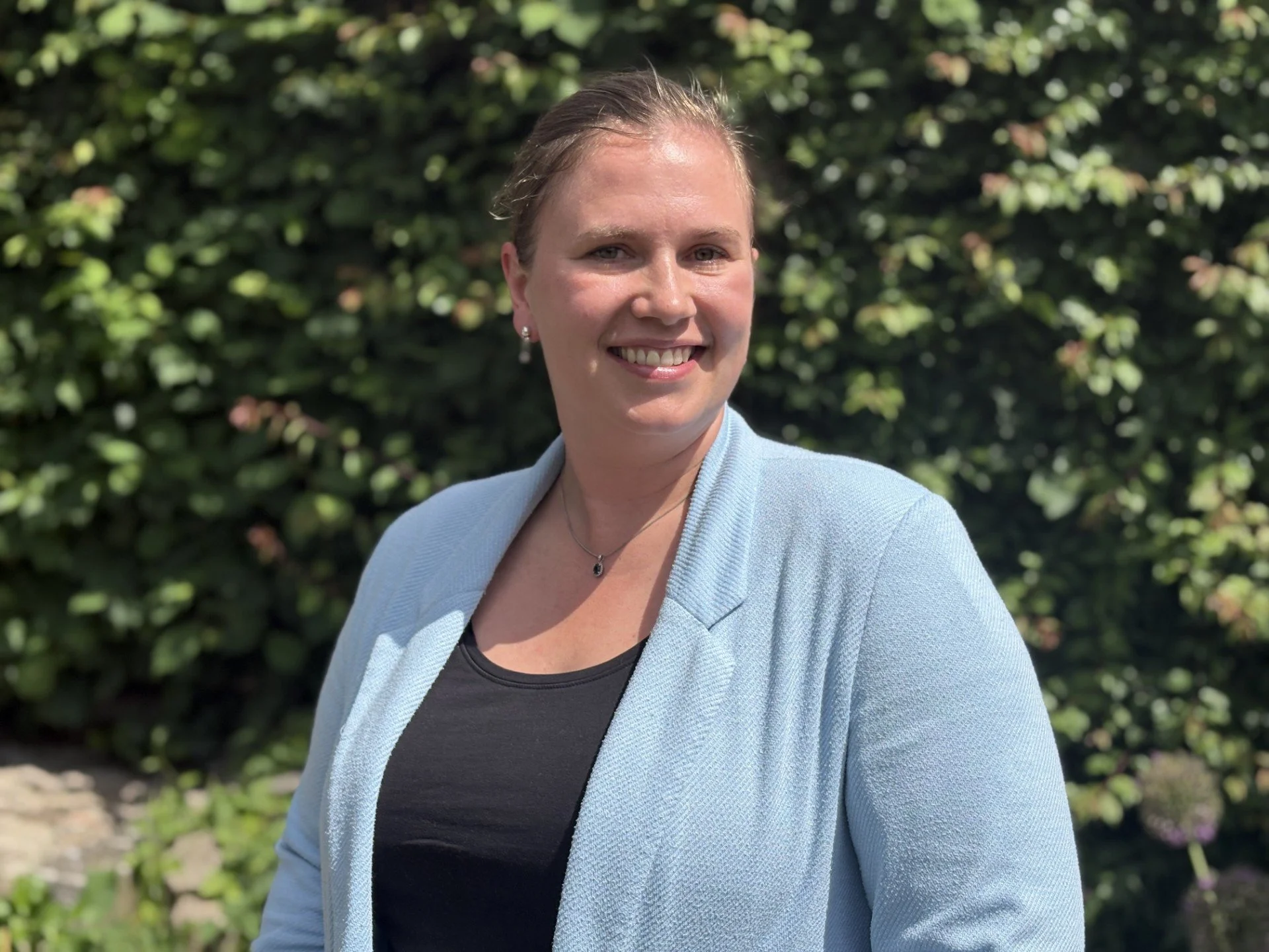 En smilende kvinde med lysebrunt hår i en lav knold, iført sorte øreringe, en lyseblå blazer og en sort top, står foran en grøn væg af buske i solsken.