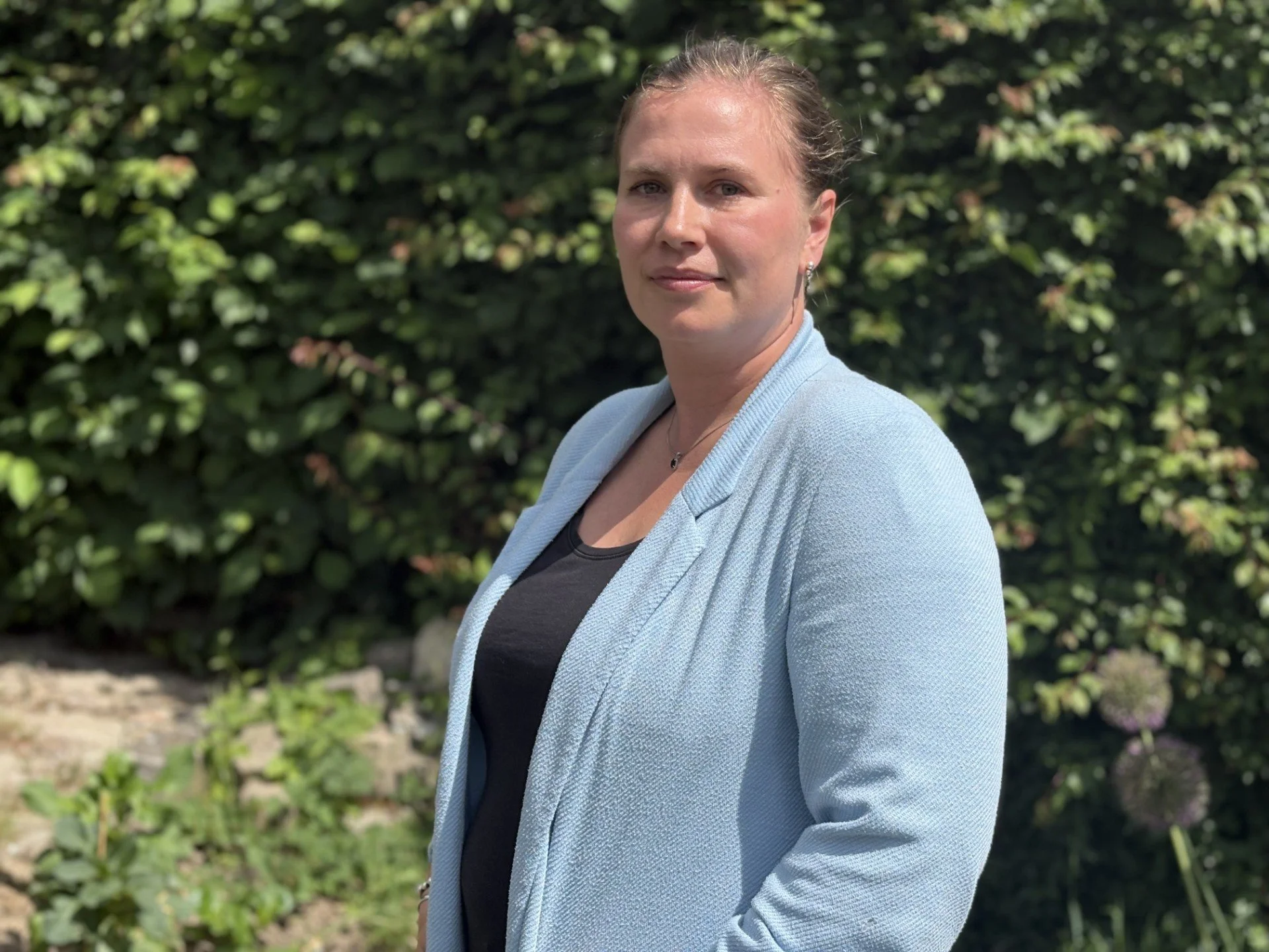 En kvinde står udenfor foran grønne buske, iført en lyseblå blazer og sort top.