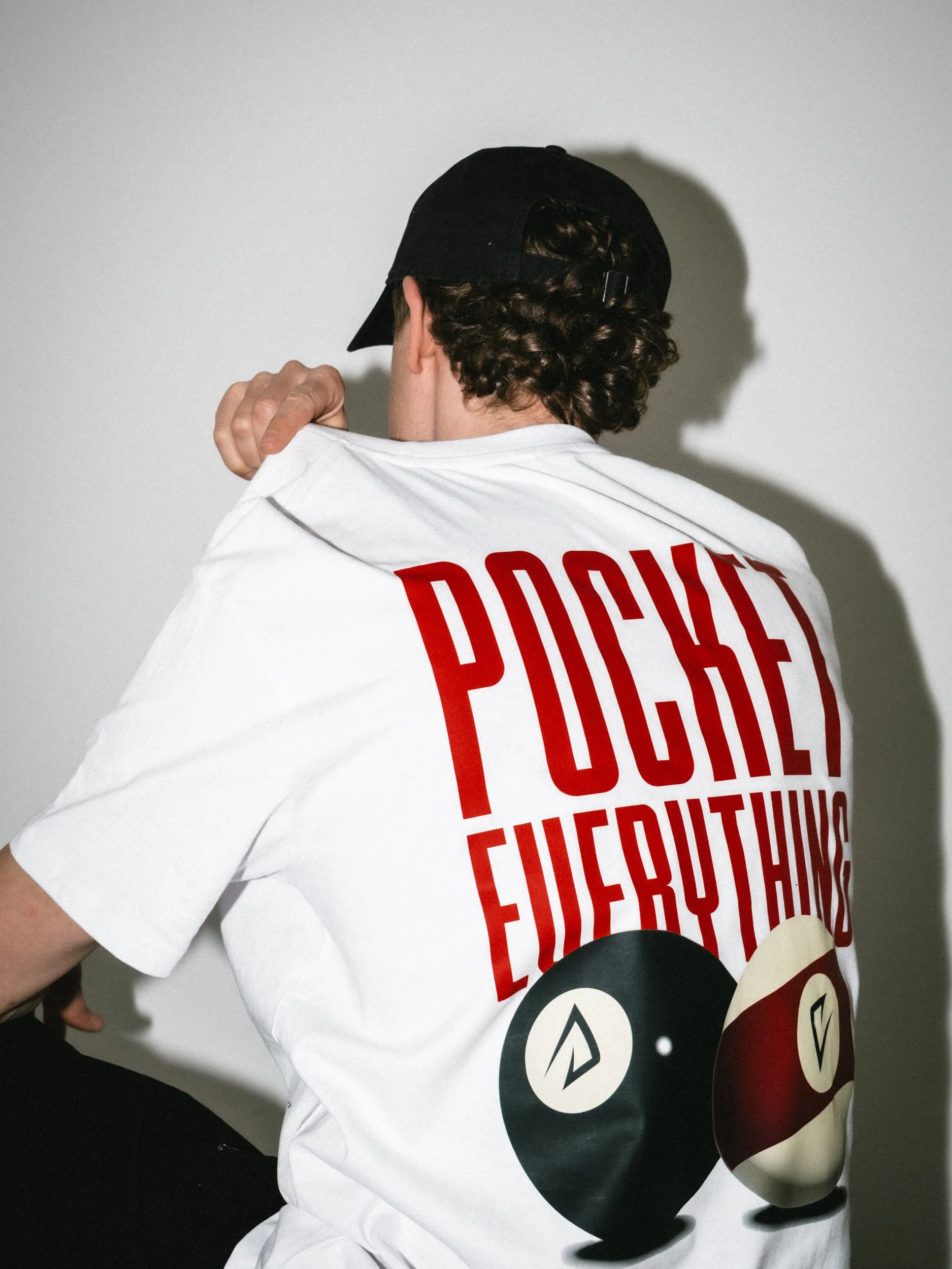 A person with curly hair and a black cap, wearing a white T-shirt with red text and graphics, is pulling the collar of the shirt over the back of the head. The shirt has the words 'POCKET? EVENINGS?' printed on it, along with yellow, red, and black circles.