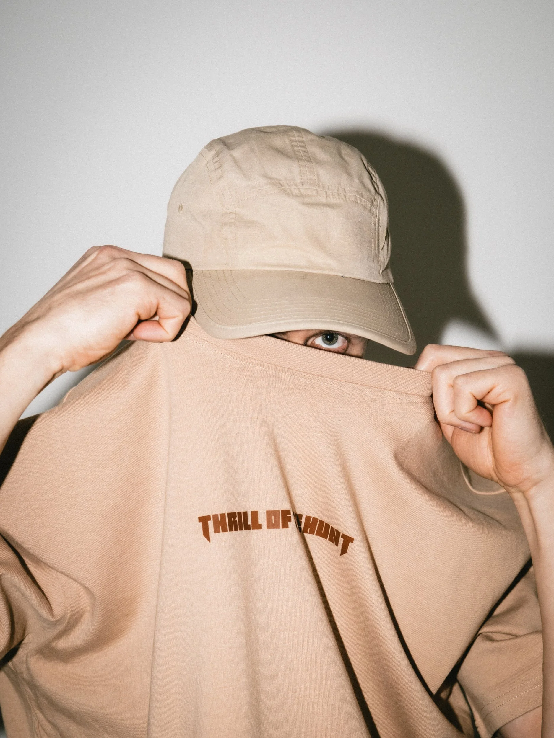 Person wearing a beige hat and beige sweatshirt with 'Thrill of Hunt' written on it, lifting the sweatshirt to partially cover their face and reveal their blue eye, against a plain background.