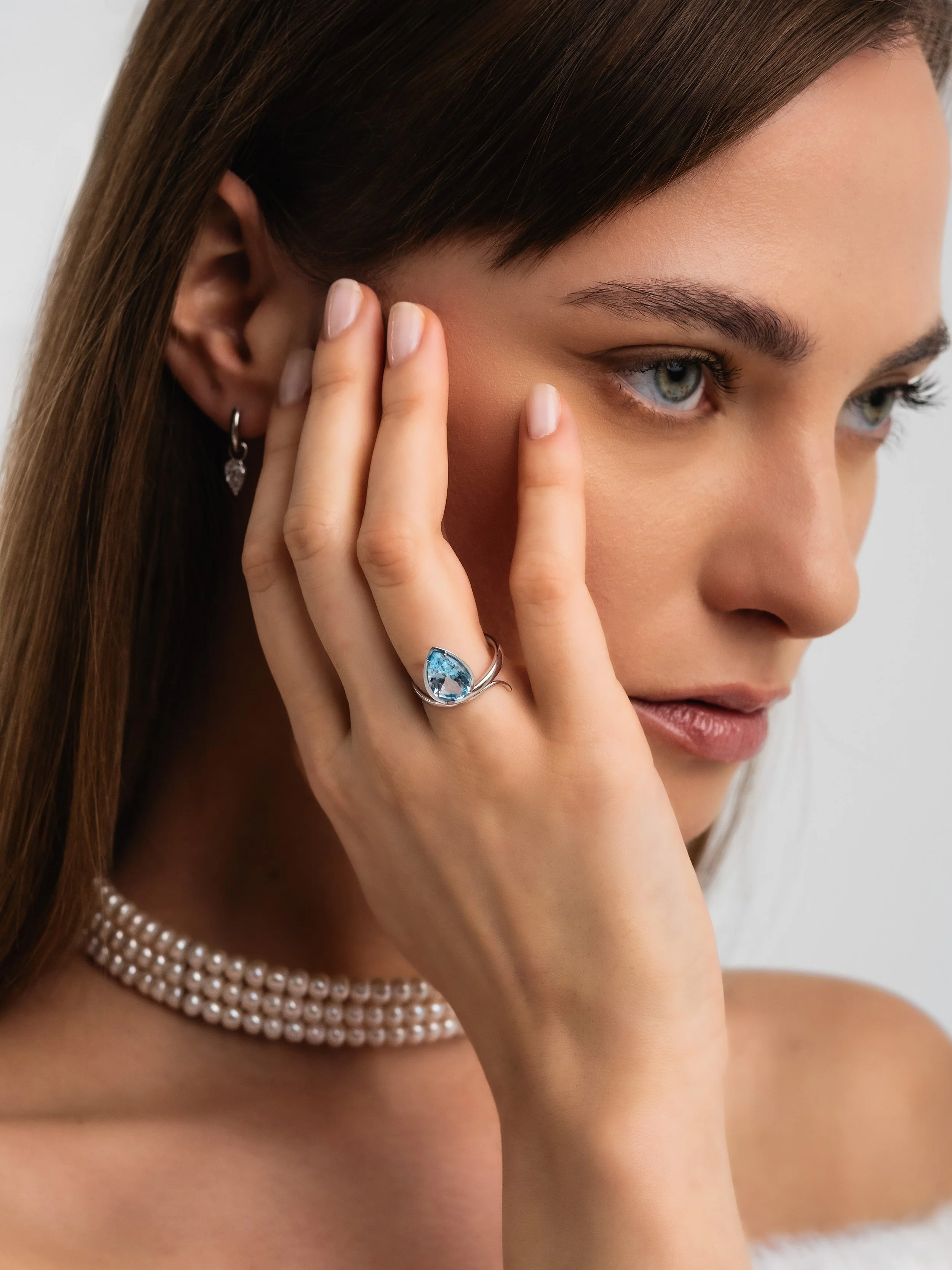 Close-up of a woman with long brown hair touching her face, wearing a pearl necklace, earrings, and a blue gemstone ring.