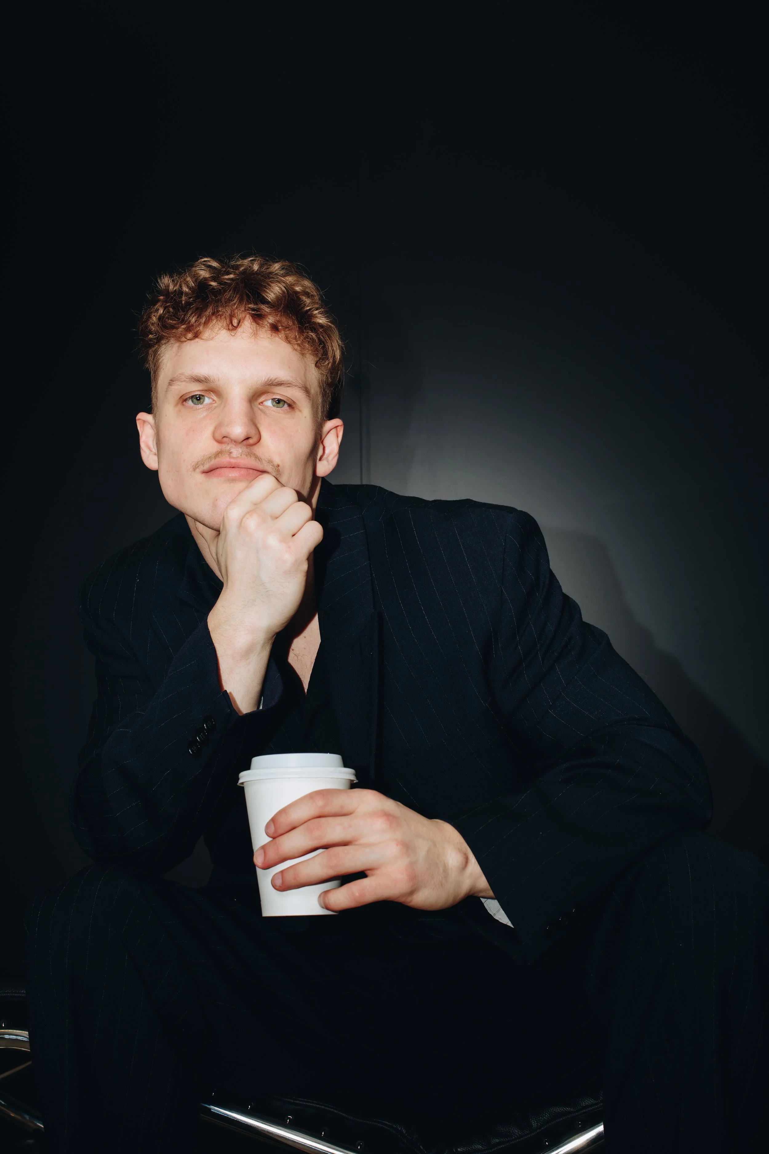 A young man with curly hair and a mustache, dressed in a dark pinstripe suit, sitting against a black background, holding a white coffee cup and resting his chin on his hand in a thoughtful pose.