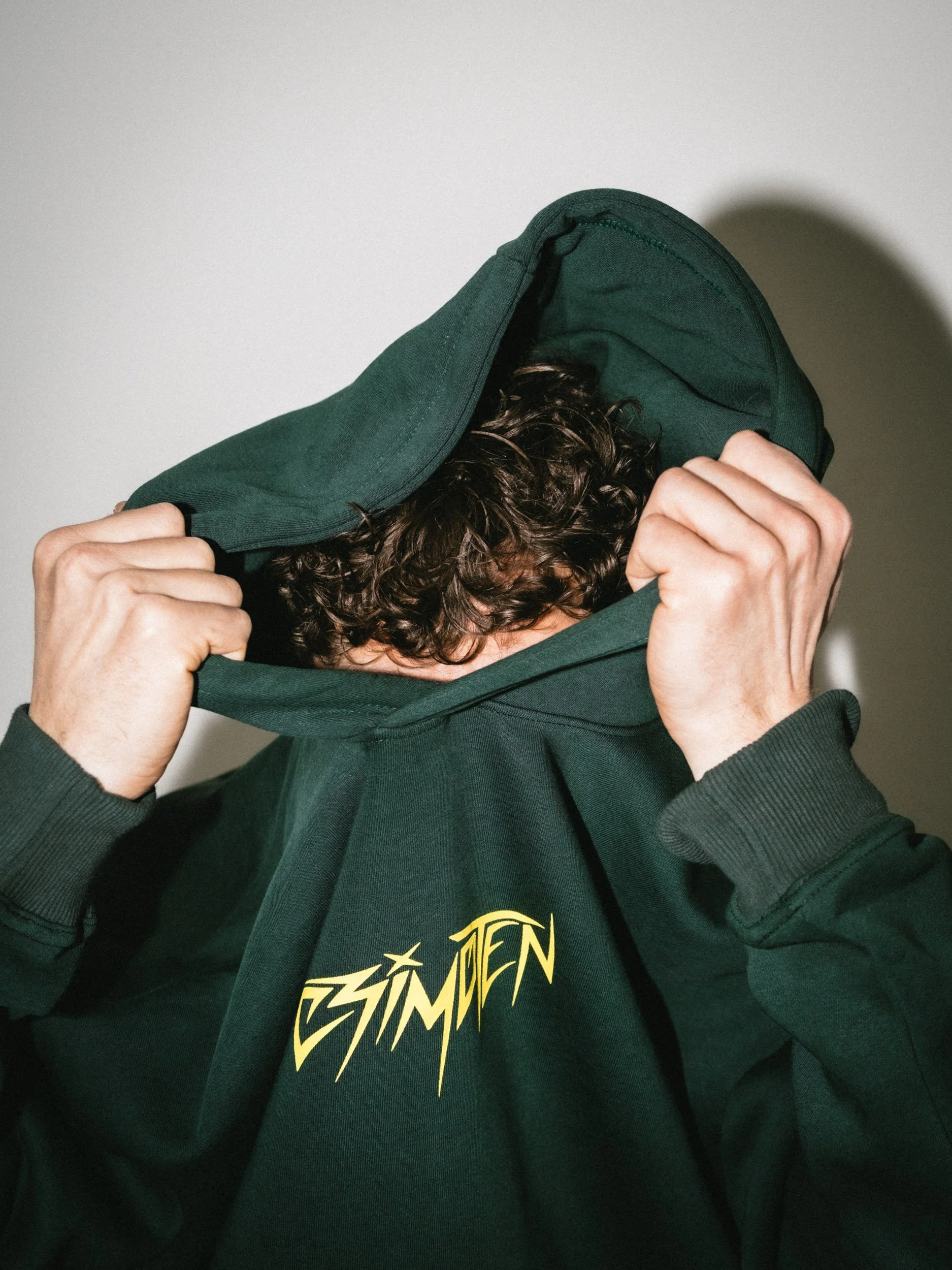 Person wearing a dark green hoodie with yellow text, pulling up the hood to cover their face.