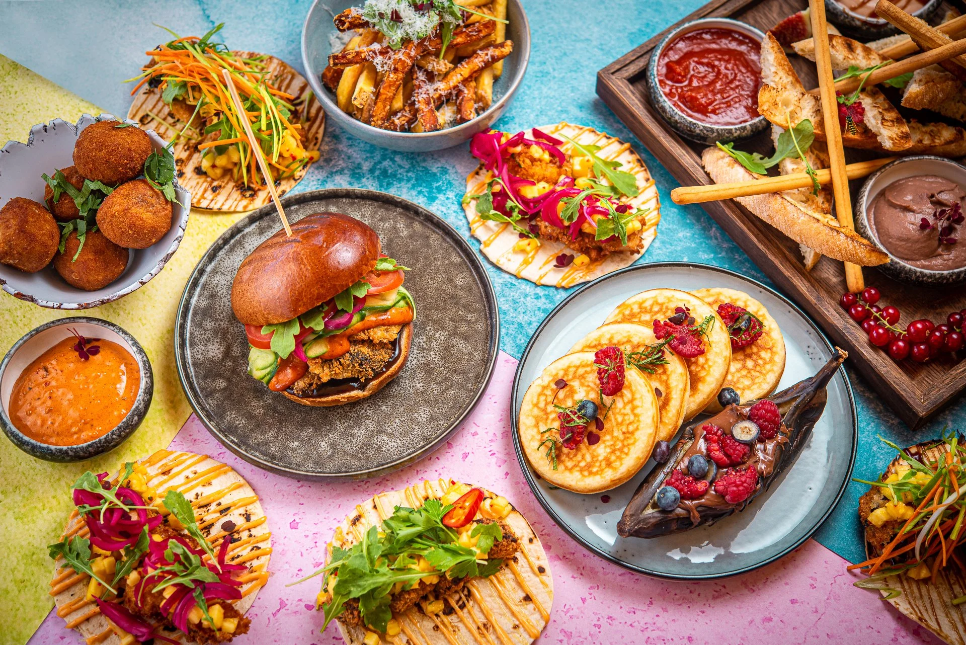 A colorful spread of various dishes including sliders, fried foods, fruit pancakes, and desserts with fresh berries, presented on vibrant colored table surfaces.