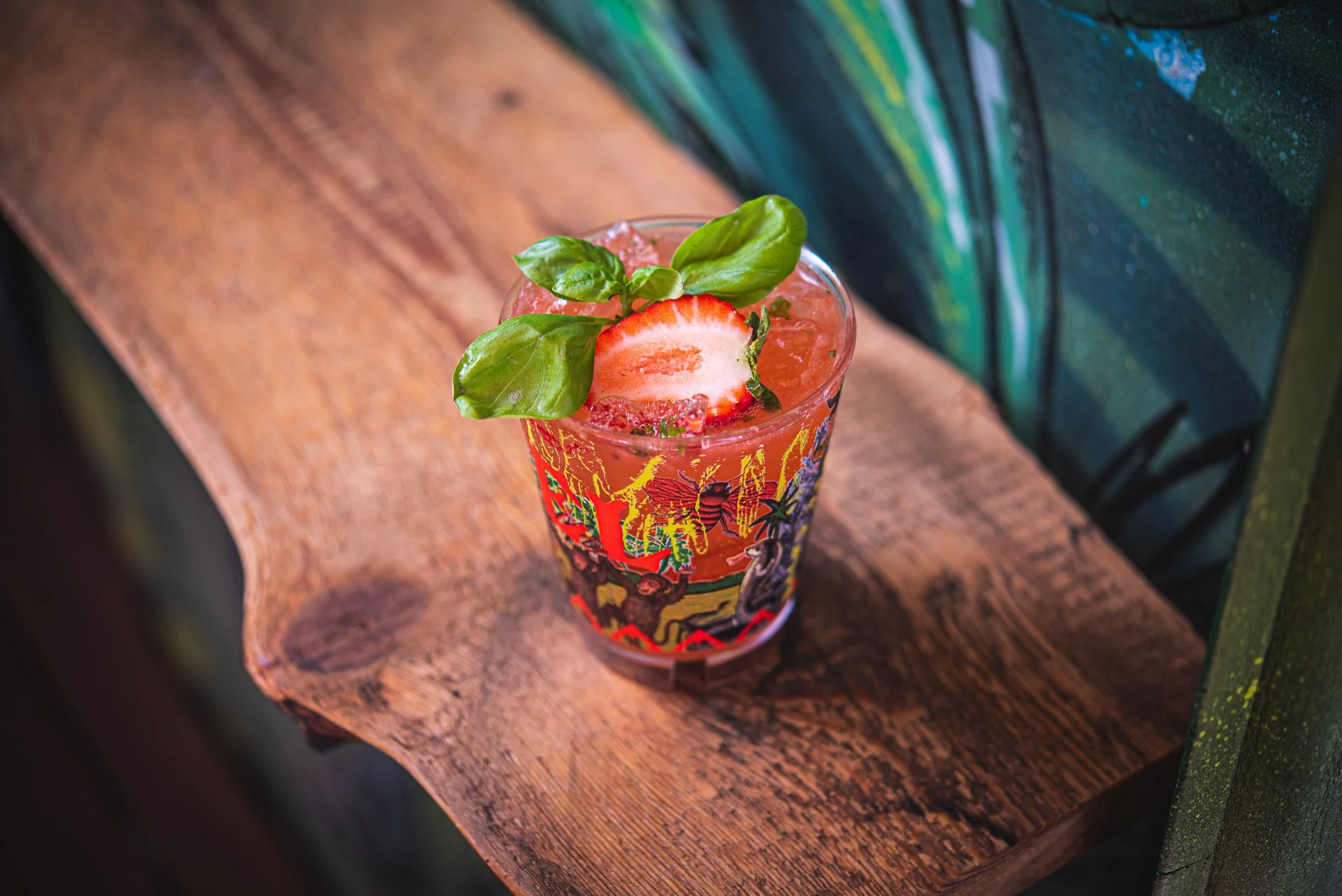 A colorful tropical cocktail garnished with a strawberry slice and fresh basil leaves, served in a glass with vibrant fruit-themed artwork on it, placed on a wooden surface.