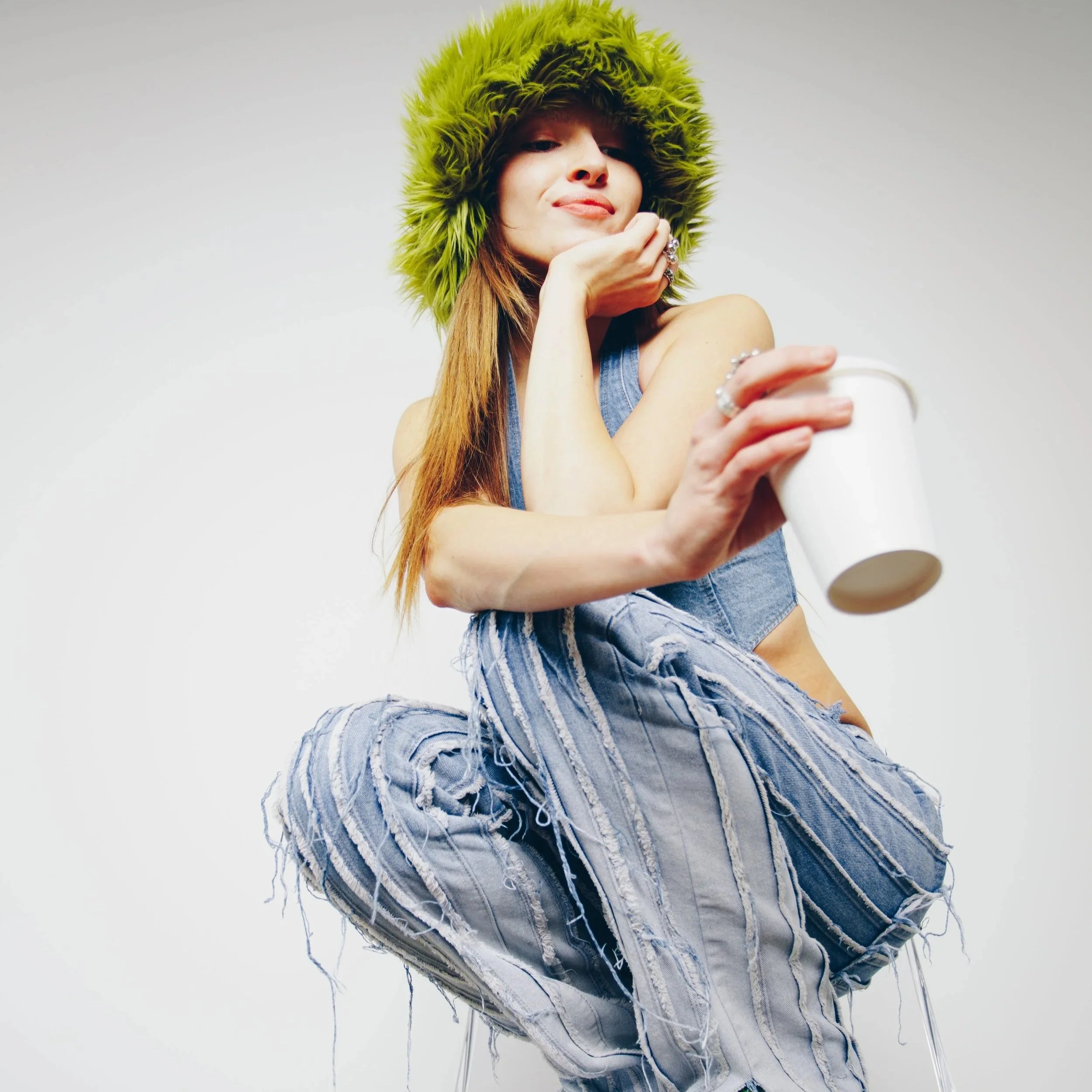 A young woman wearing a lime green furry hat and distressed denim overalls, sitting with her knees up and holding a white cup, posing against a plain white background.