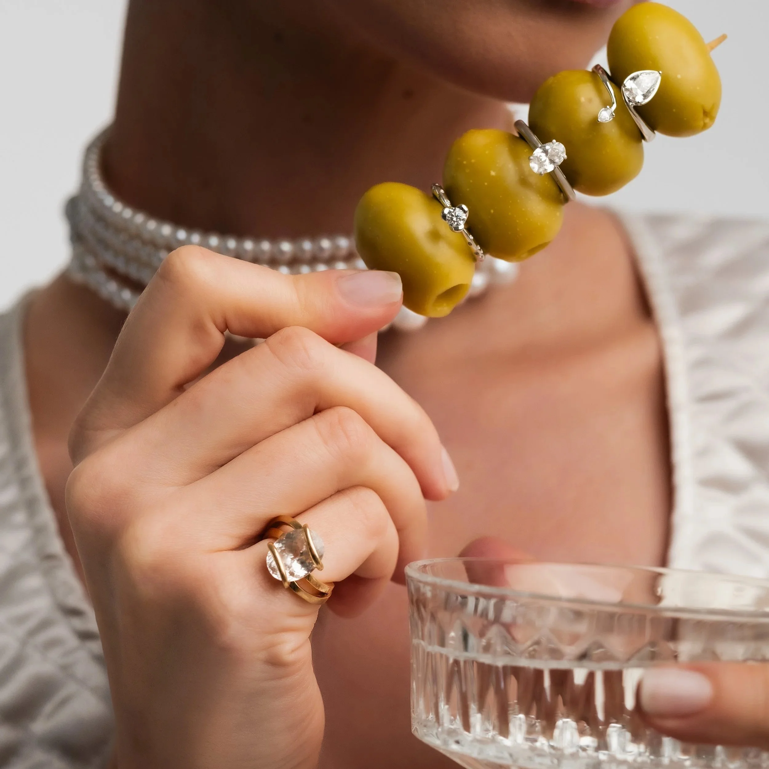 Person wearing pearls, rings with diamonds, holding a glass of water and a skewer with green olives that have rings and earrings on them.