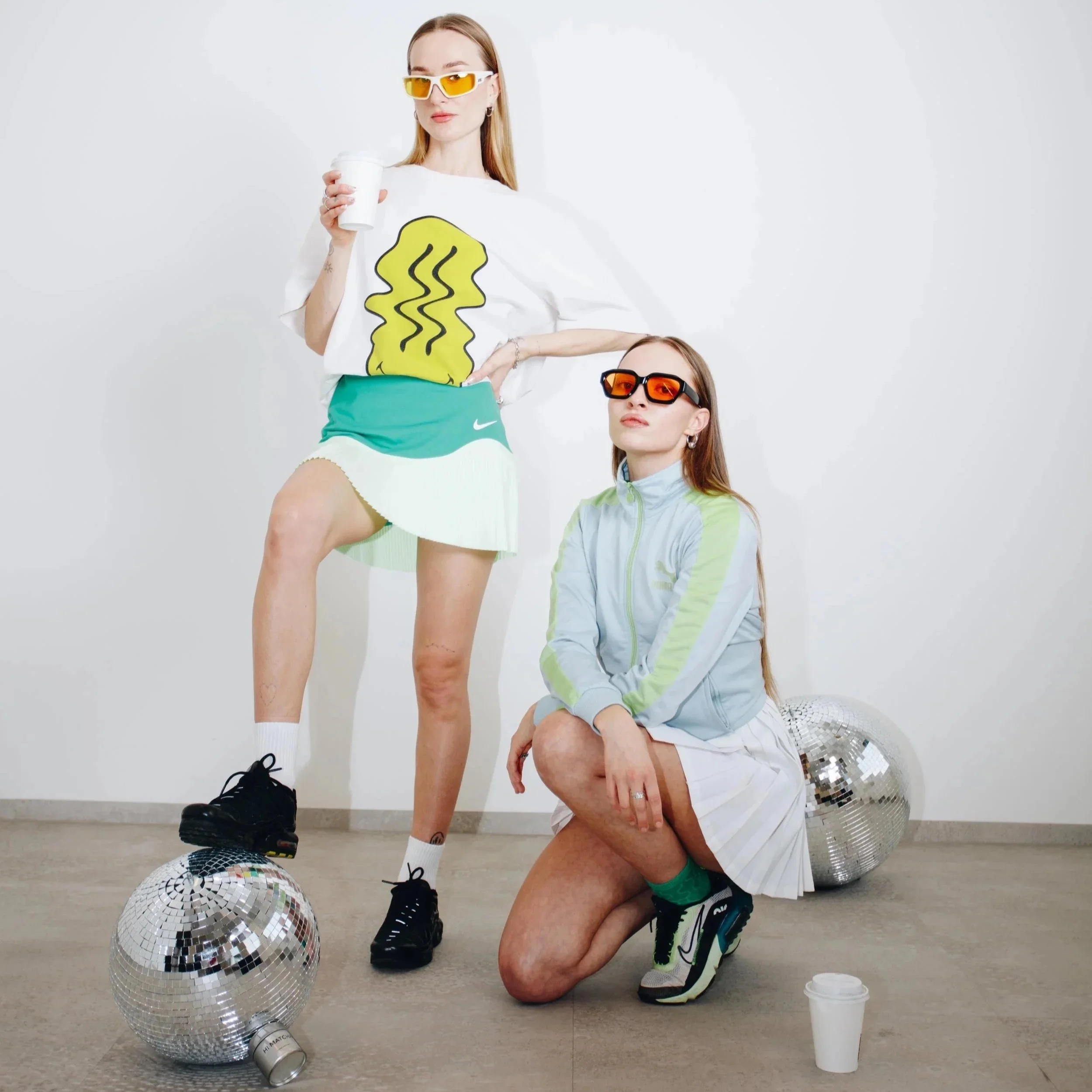 Two young women in athletic clothing with colorful sunglasses, posing with disco balls in a minimalist setting. One is standing with a coffee cup, the other kneeling.