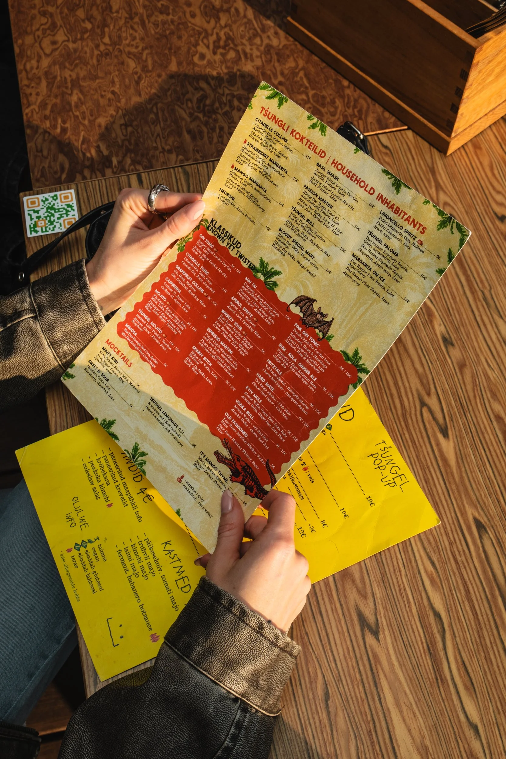 Person holding a colorful menu with drinks and food options, sitting at a wooden table in a restaurant, with a yellow sheet of paper underneath.