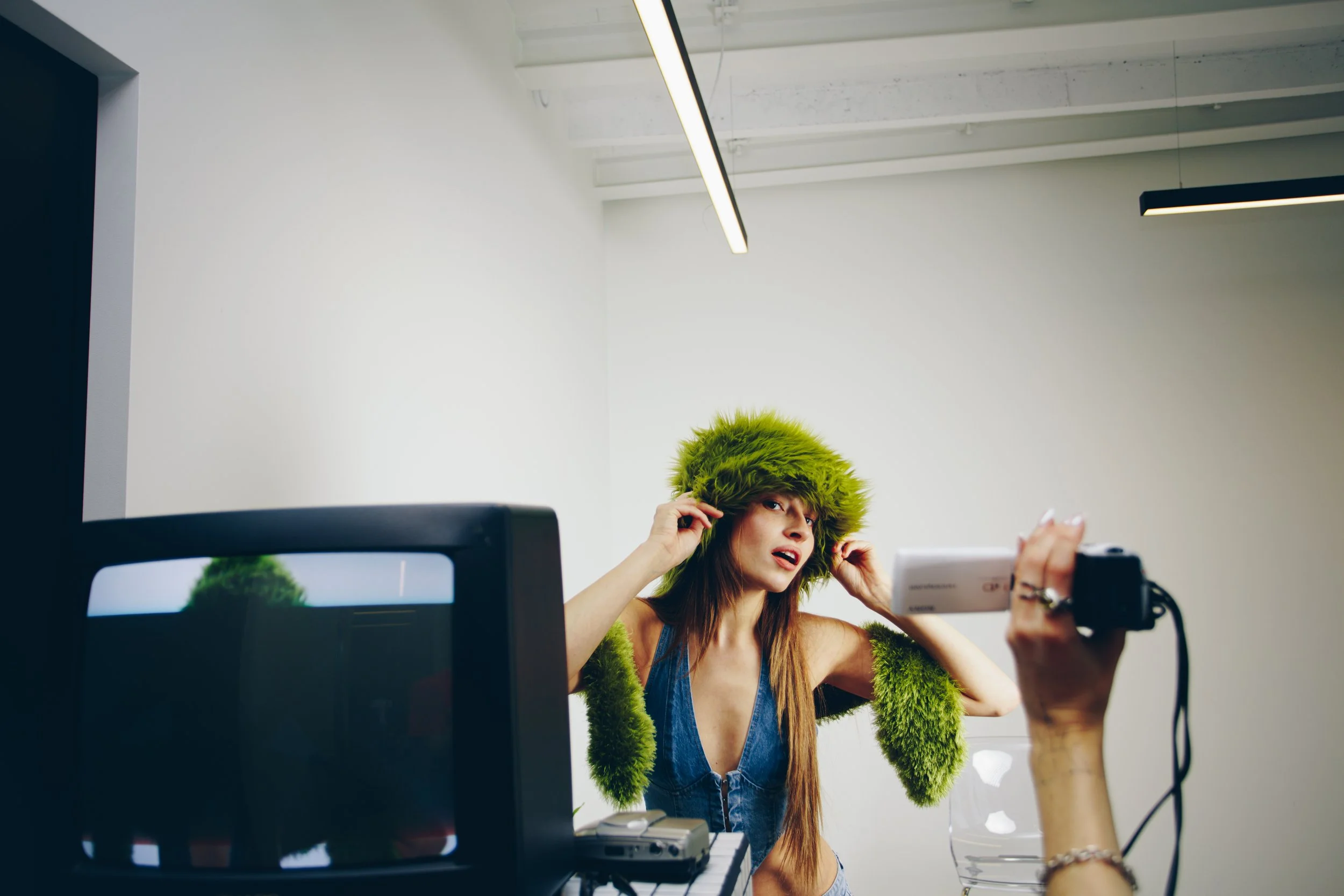 A woman wearing a green furry hat and matching green furry sleeves being photographed in a studio with a white wall background and a monitor in the foreground.