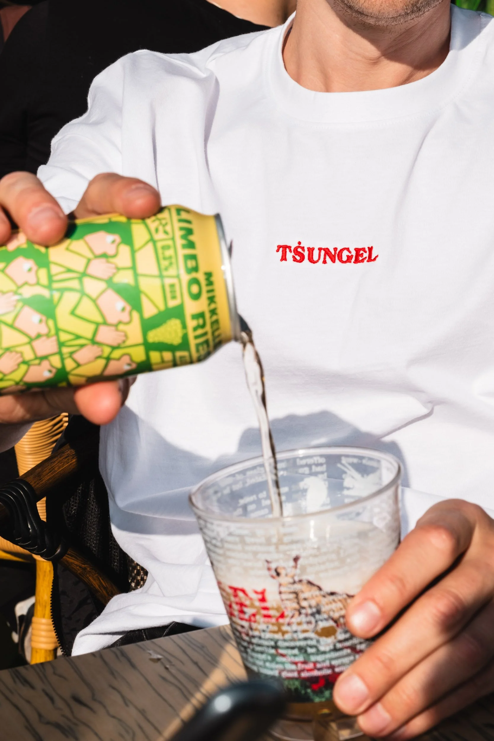 A person in a white shirt with red embroidery reading "TSUNGEL" is pouring a light-colored beverage from a green can into a glass with a deer illustration on it. Another person in a black shirt is partially visible behind.