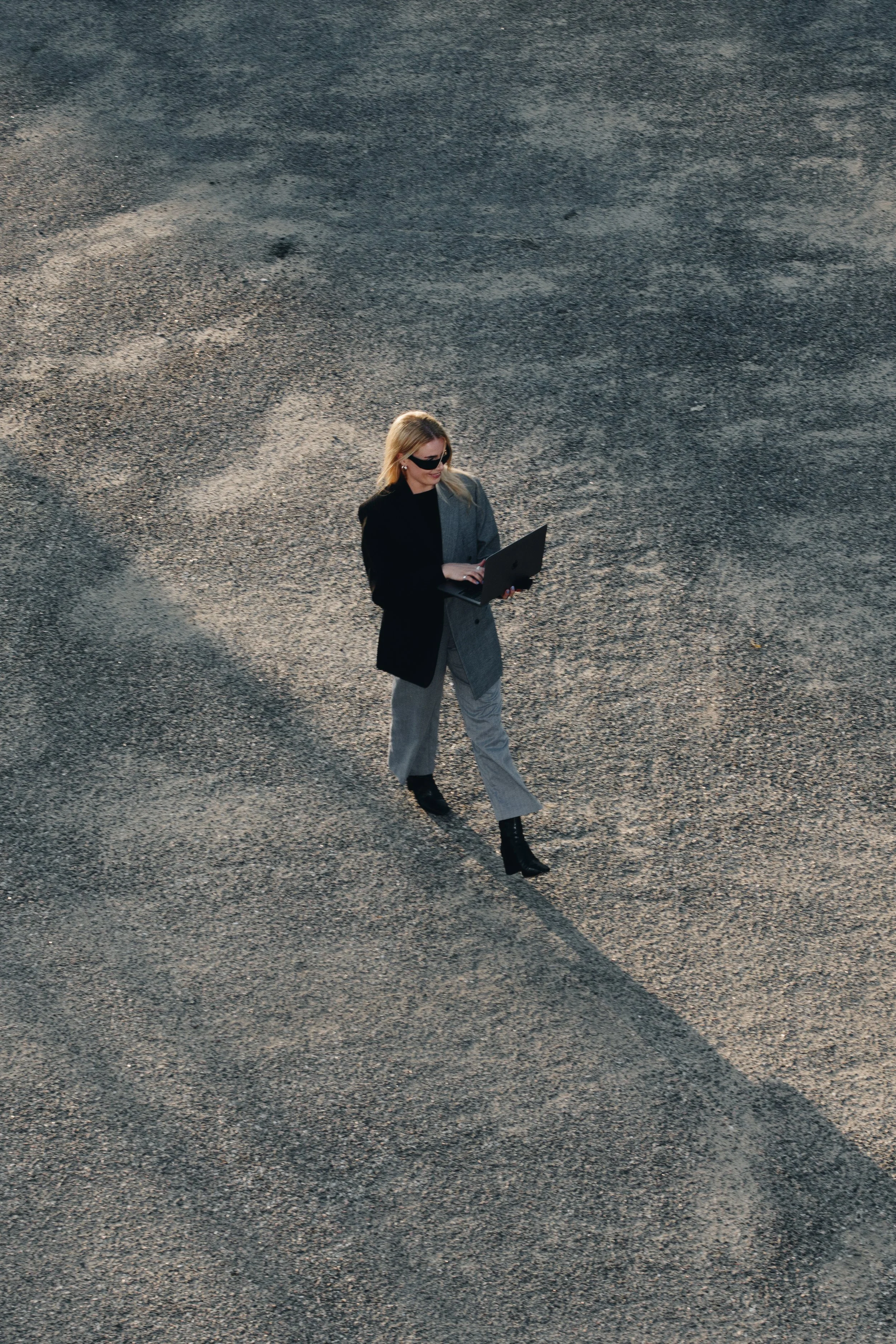 A woman wearing sunglasses and business attire walking on a gravel surface while working on a laptop.